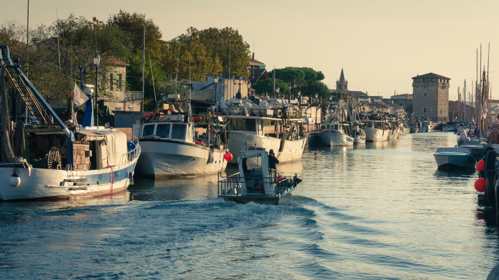 View of the port, with numerous fishing boats moored along the canal and a small motorboat navigating through the water