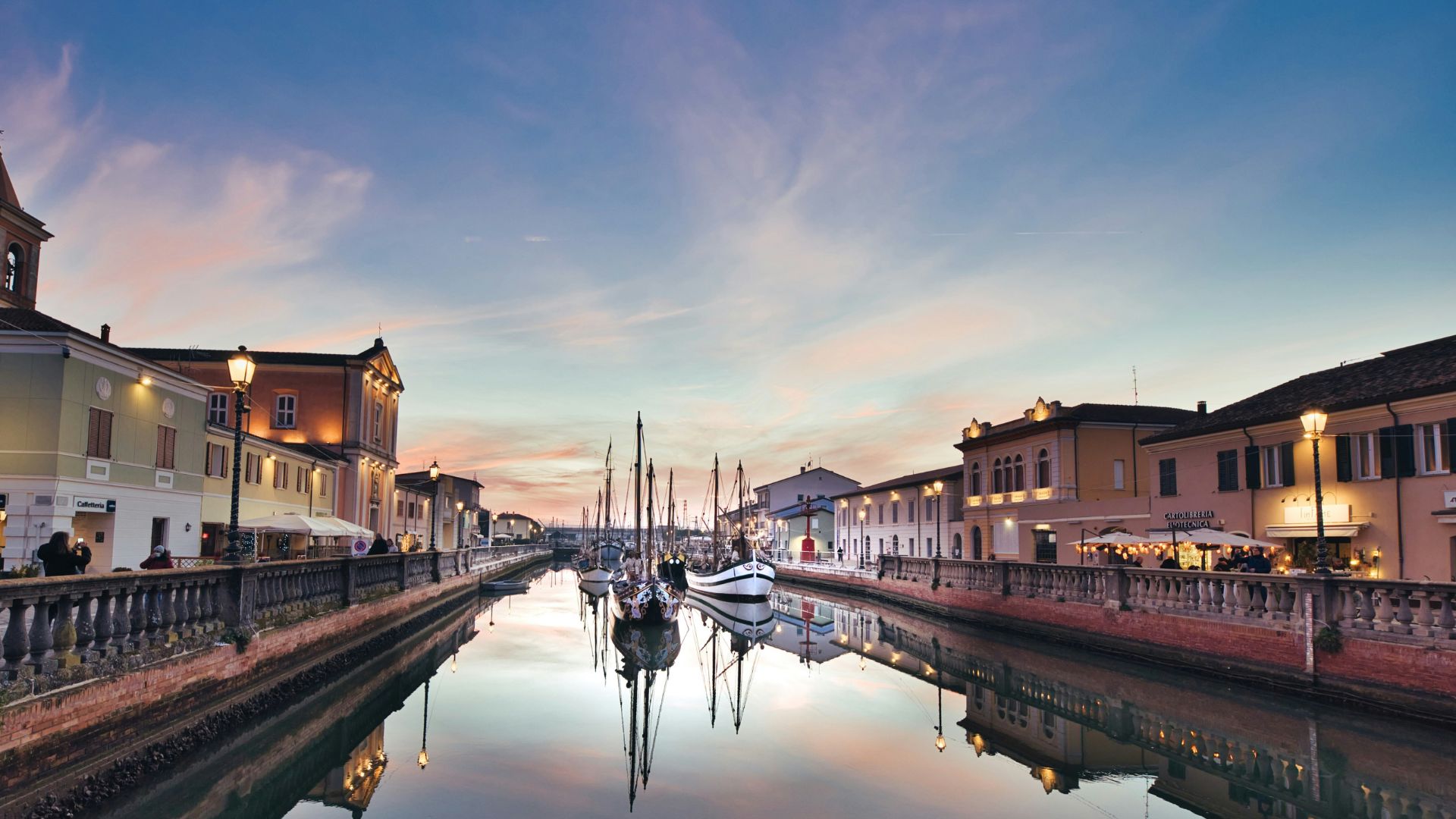 Porto Canale Leonardesco in Cesenatico, Italy at dusk, with buildings lining the canal and boats moored in the center, all reflected in the calm water. The sky is a soft blend of pink and blue, and the scene is lit by street lamps and building lights.