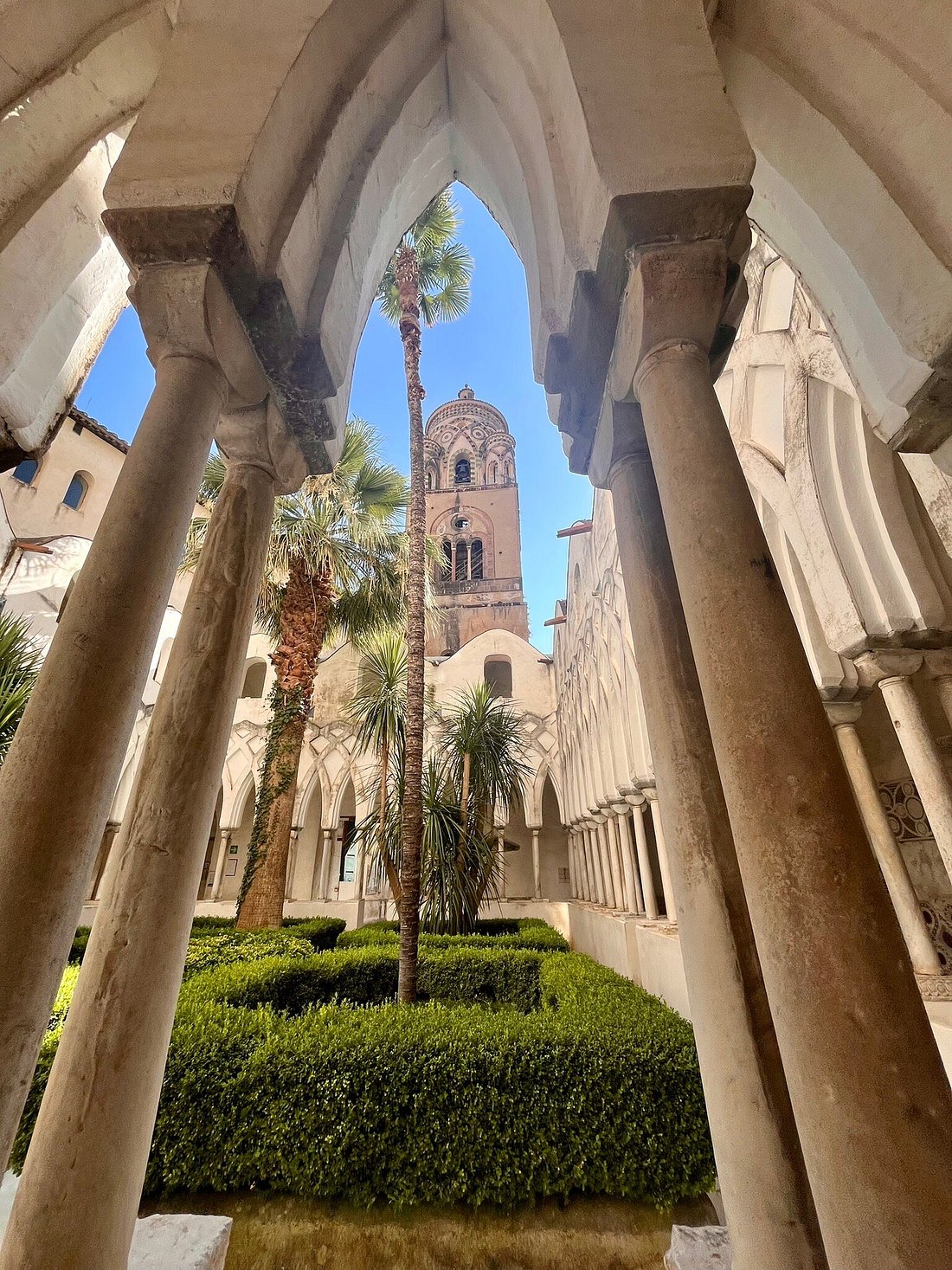 View of Chiostro del Paradiso courtyard framed by arched columns, with palm trees and a bell tower in the background.