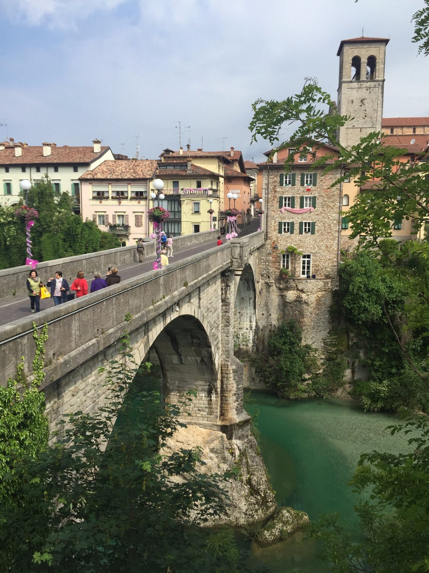 Scenic view of Cividale del Friuli, Italy, with people walking across the historic stone Ponte del Diavolo bridge over emerald-green water, surrounded by charming colorful buildings and a bell tower.