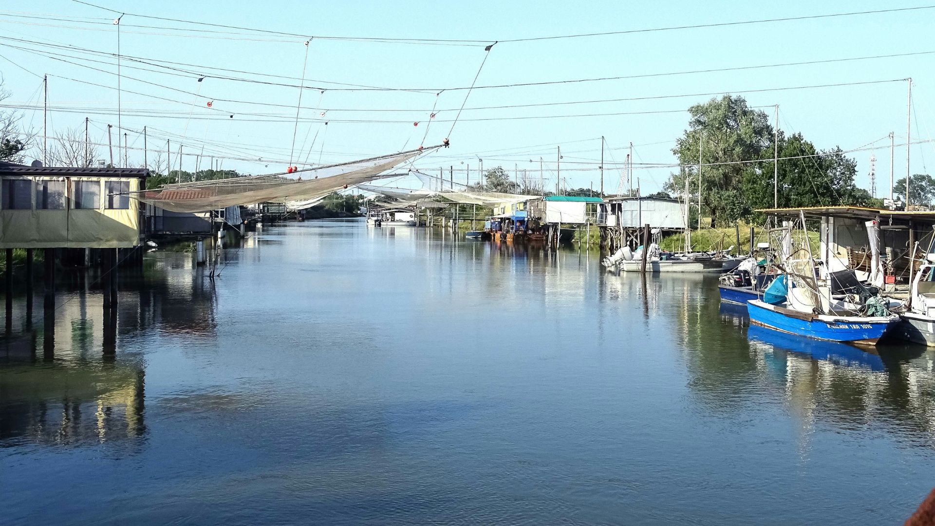 The image shows a view of Comacchio lagoons, characterized by fishing huts on stilts and traditional fishing nets and includes several small boats moored along the waterways.