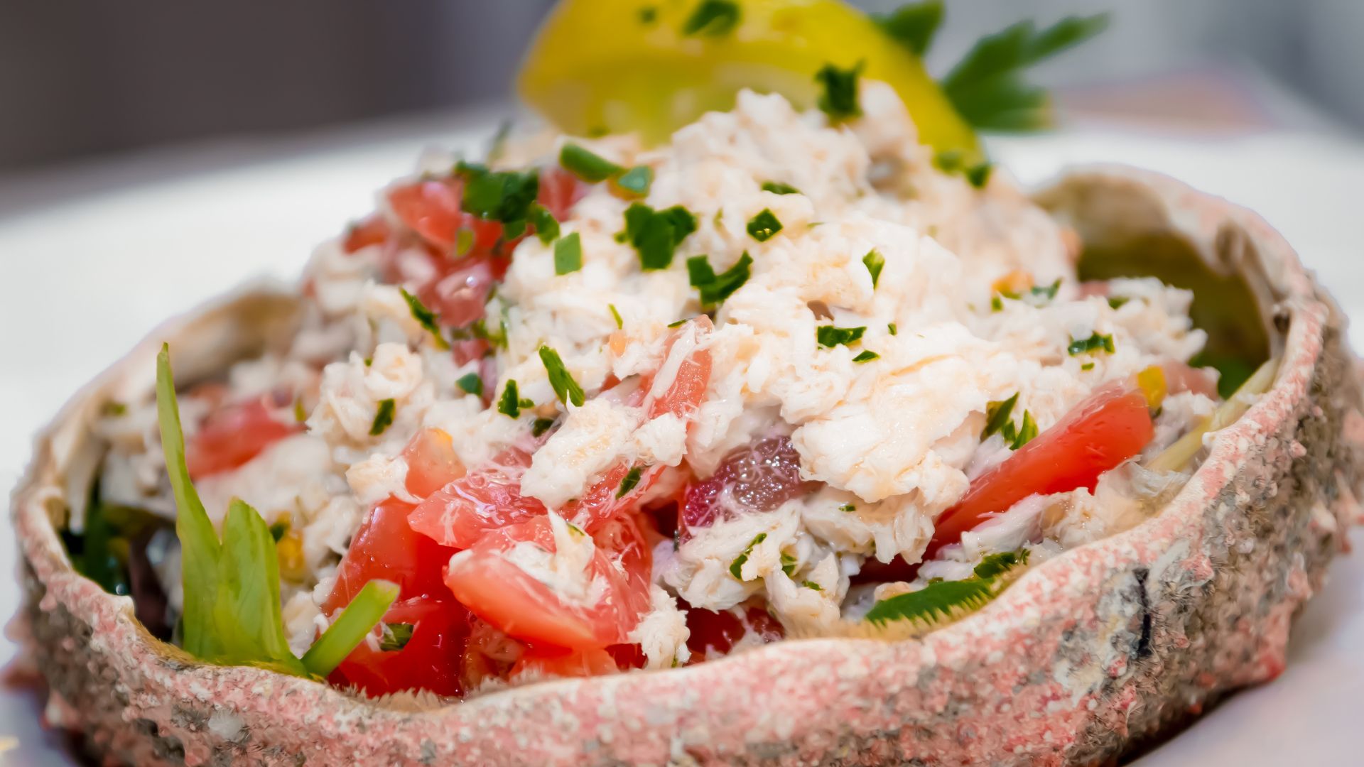 The image shows a crab salad served in a crab shell. The salad appears to contain crab meat, tomatoes, and herbs, and is garnished with a lemon wedge. 