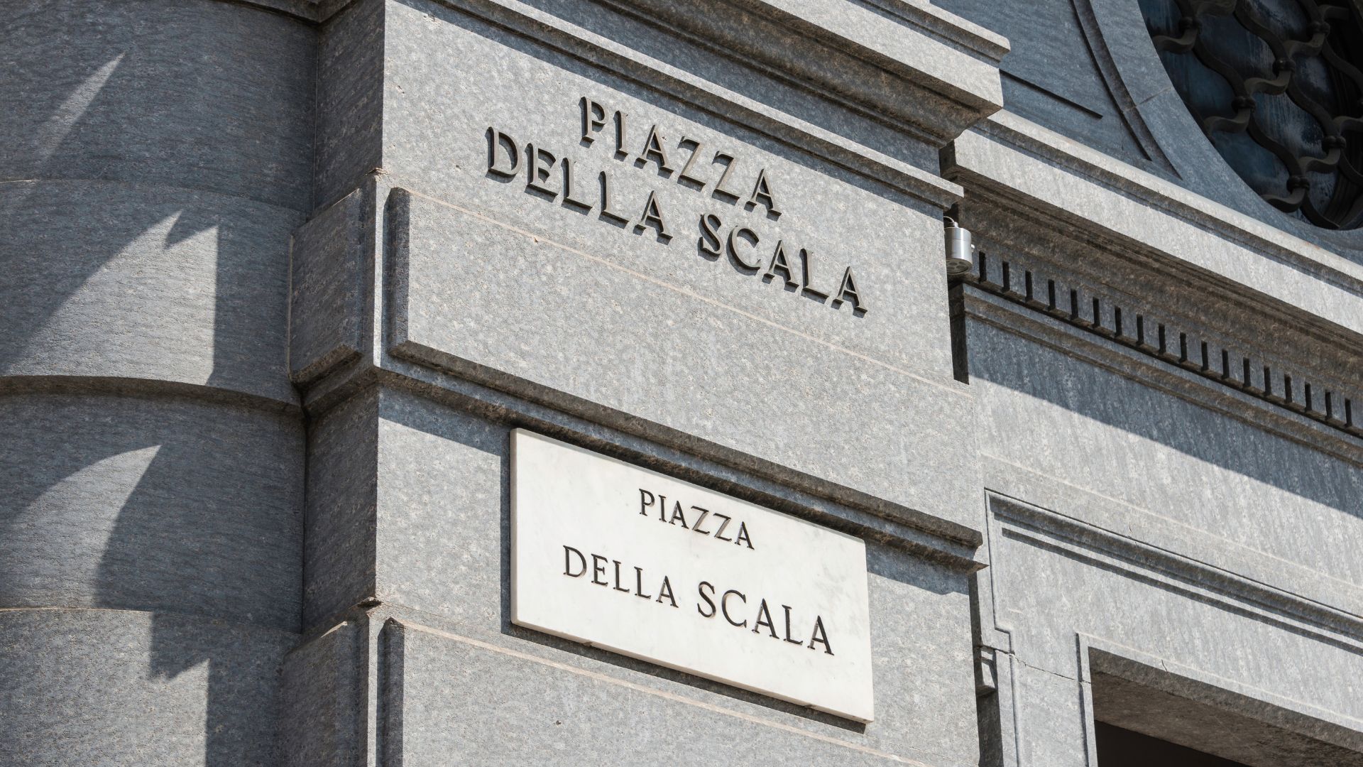 A close-up view of a building corner displays two signs, one engraved directly into the stone and the other a plaque, both reading "Piazza Della Scala." The signs mark the location of Piazza della Scala, a famous square in Milan, Italy, known for the Teatro alla Scala opera house and Palazzo Marino.
