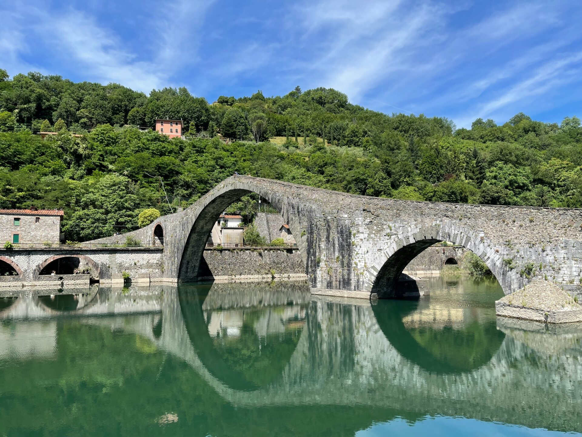 Stone arch Devil’s Bridge in Cividale del Friuli, Italy, reflected in the calm green river below, surrounded by lush hills and historic buildings.