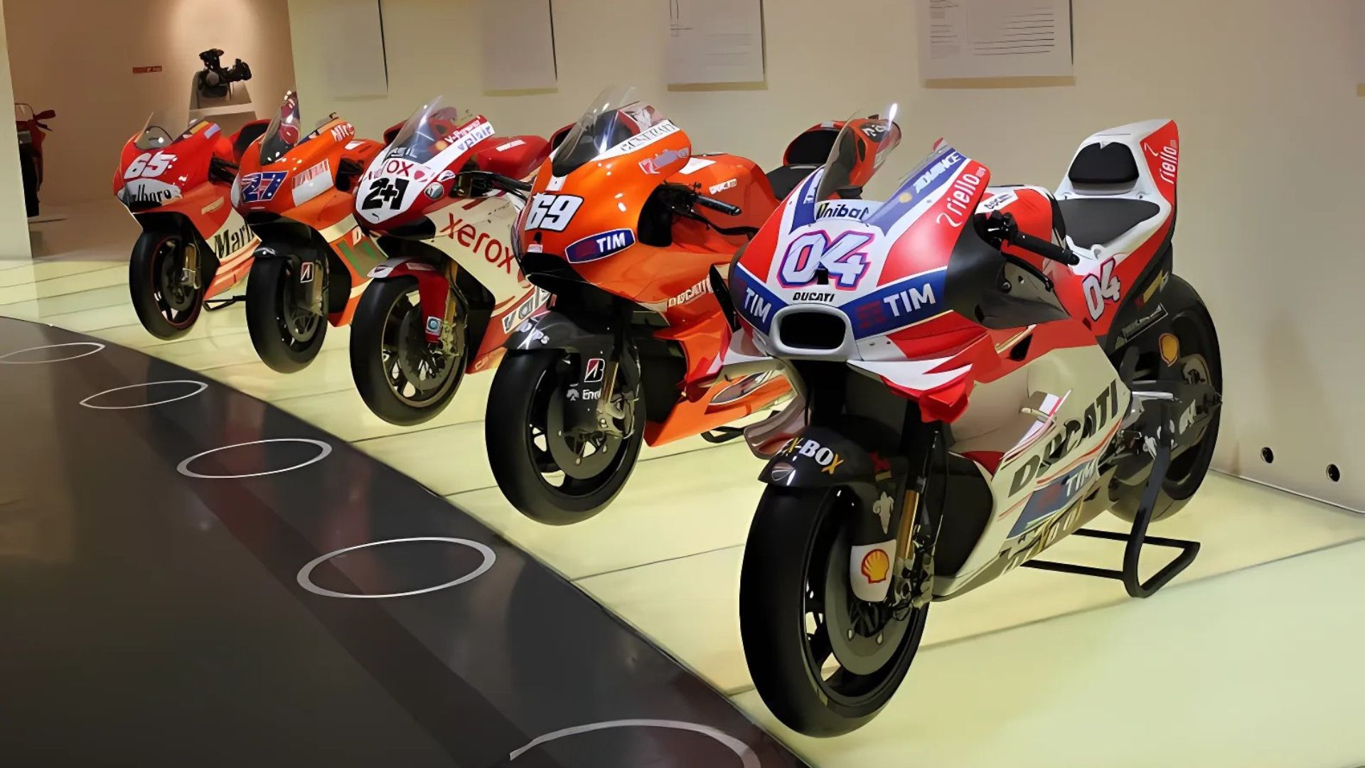 A lineup of Ducati Desmosedici motorcycles is displayed at the Ducati Museum, showcasing their evolution with different racing liveries and sponsor logos, including "TIM," "Xerox," and "Unibat.