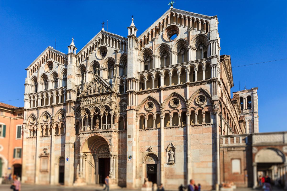 The image shows the Ferrara Cathedral. The facade features intricate carvings, arched windows, and decorative elements characteristic of the period.