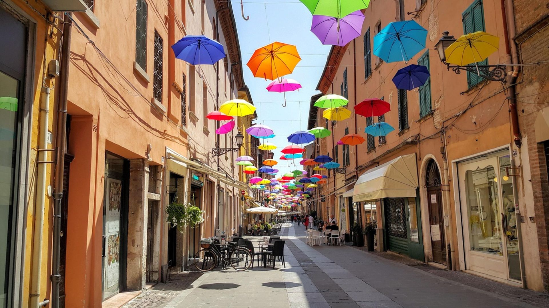The image shows a street in Ferrara, Italy, adorned with colorful umbrellas suspended overhead. The street is paved with stone and has a bicycle parked on the side, adding to the scene's charm.