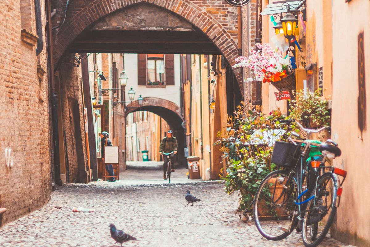 Image of Via delle Volte, Ferrara, Italy (part of the former Jewish Ghetto) showing its medieval arched passageways. Bicycle on the right side, some birds on the passageway and some 2 people at the background. 