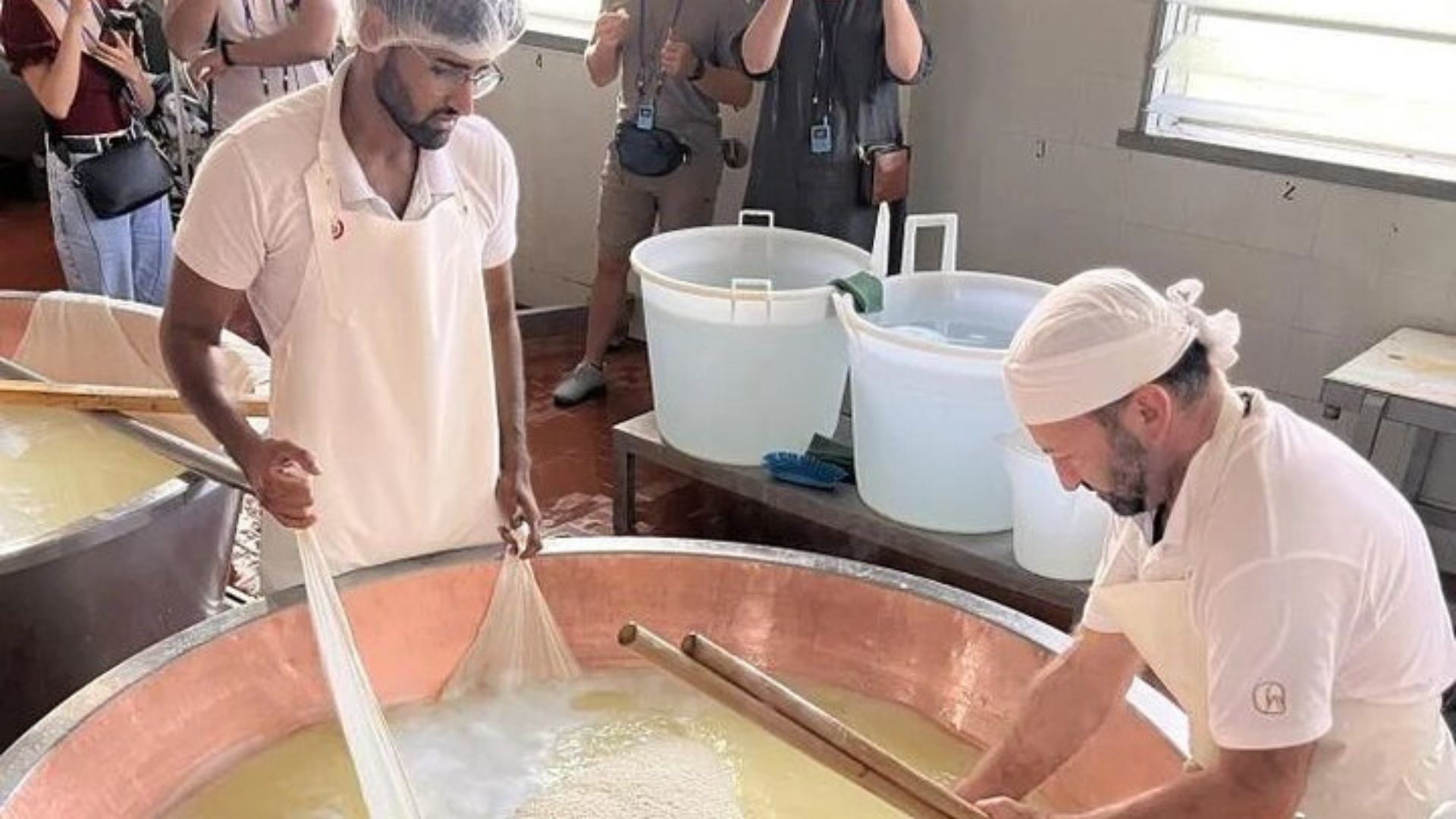 Two cheesemakers work in a copper vat making Parmigiano Reggiano. They are wearing white uniforms and hairnets, and several tourists observe the process in the background. The vat is filled with a milky liquid, and one cheesemaker uses a cloth to gather solids while the other uses a wooden tool to stir or manipulate the contents.
