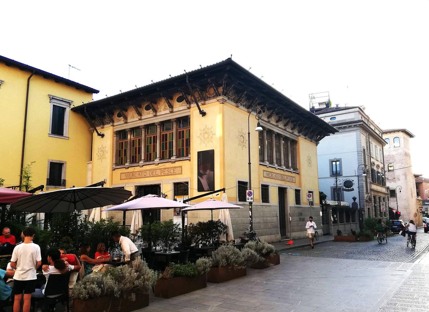 Exterior of Galleria Tina Modotti in Udine, Italy, a historic yellow building with decorative wooden eaves and large windows, with people dining under umbrellas in a cozy outdoor café setting.