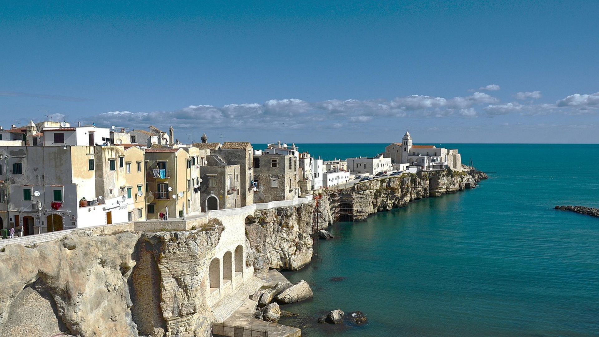 A coastal view of Gargano National Park featuring a rocky cliff with lush greenery, a small boat in the clear blue water, and the horizon of the sea under a light sky.