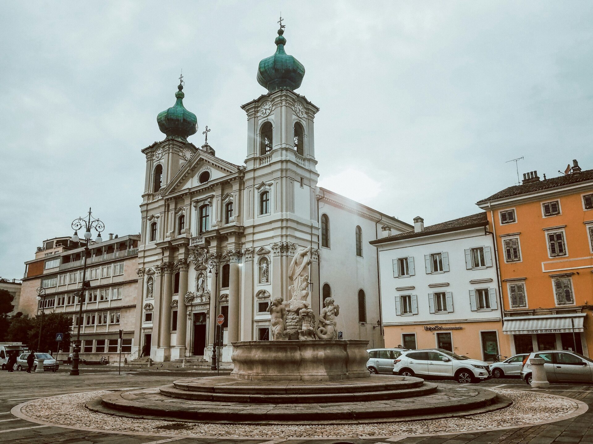 Historic church with twin green domes and a grand fountain in front, located in Gorizia, Province of Gorizia, Italy, surrounded by colorful buildings and parked cars on a cloudy day.