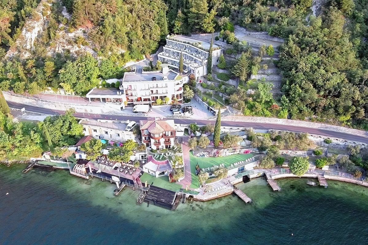Aerial view of Surf Hotel Pier on Lake Garda, featuring multiple buildings, a waterfront area with docks, and surrounding greenery. The hotel complex includes various structures, from the main hotel buildings to lakeside facilities, set against a backdrop of lush trees and a winding road.
