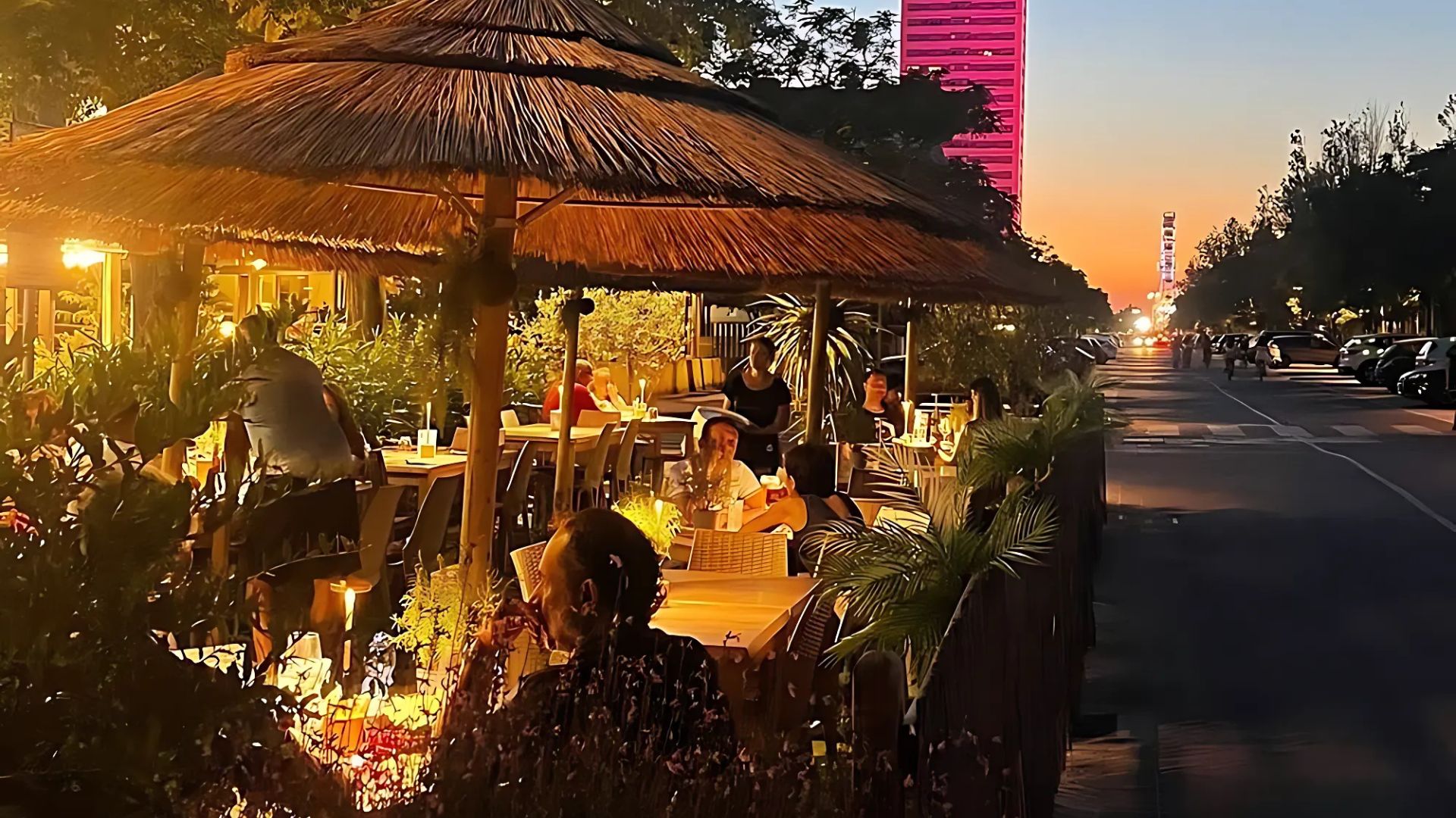 Outdoor Jam Session restaurant in Cesenatico seating area with thatched umbrellas at dusk, with a brightly lit pink skyscraper in the background.