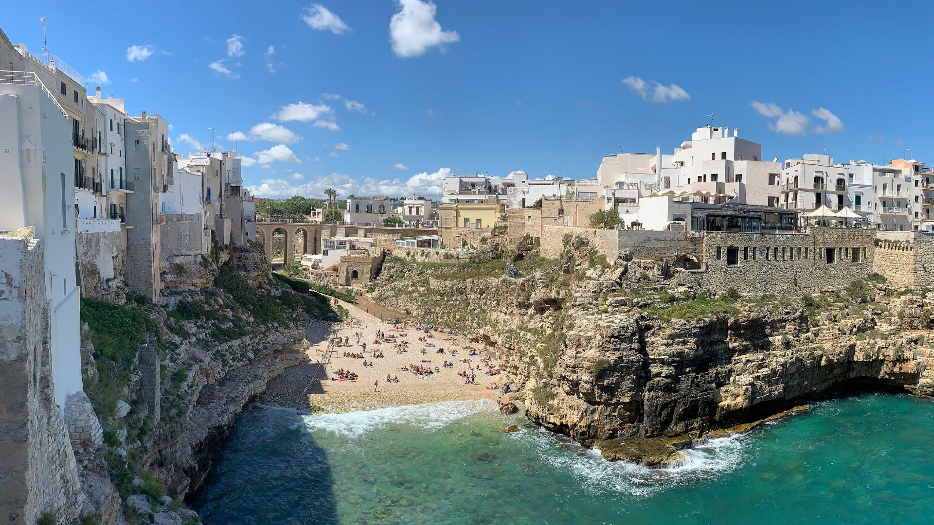 Lama Monachile beach in Polignano a Mare, Italy, is nestled between rocky cliffs and white buildings, with turquoise waters and a sandy shore dotted with people.