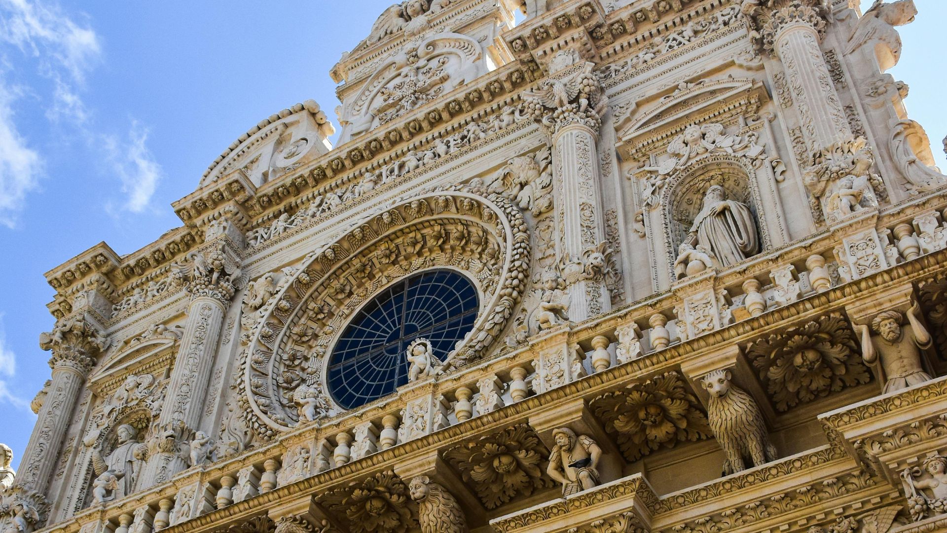 Close-up of an ornate, round window with a grid pattern, surrounded by detailed carvings of angels and stone flower floral motifs,  of the Basilica di Santa Croce in Lecce.Close-up of an ornate, round window with a grid pattern, surrounded by detailed carvings of angels and stone flower floral motifs,  of the Basilica di Santa Croce in Lecce.