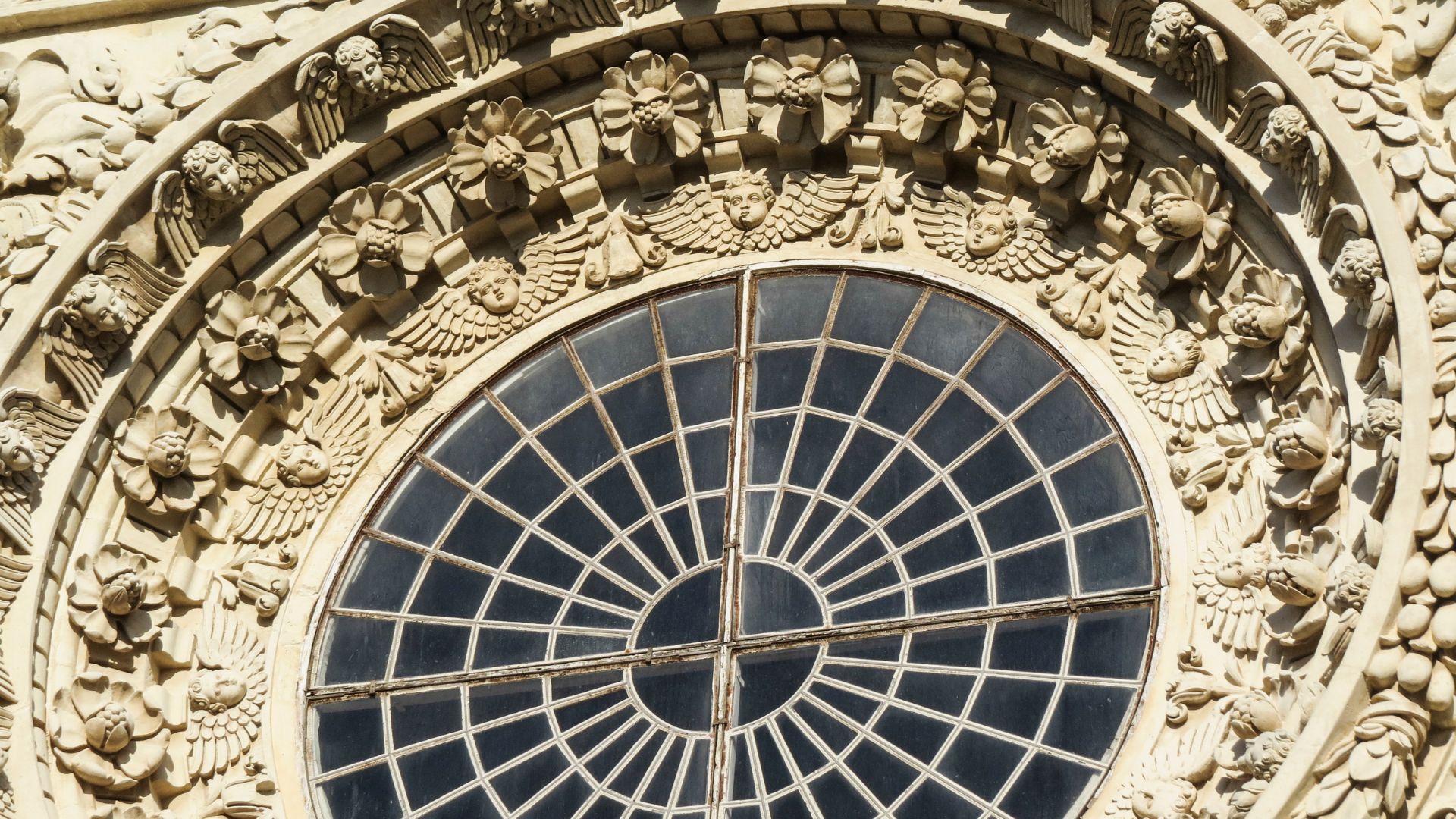 Close-up of an ornate, round window with a grid pattern, surrounded by detailed carvings of angels and stone flower floral motifs,  of the Basilica di Santa Croce in Lecce.