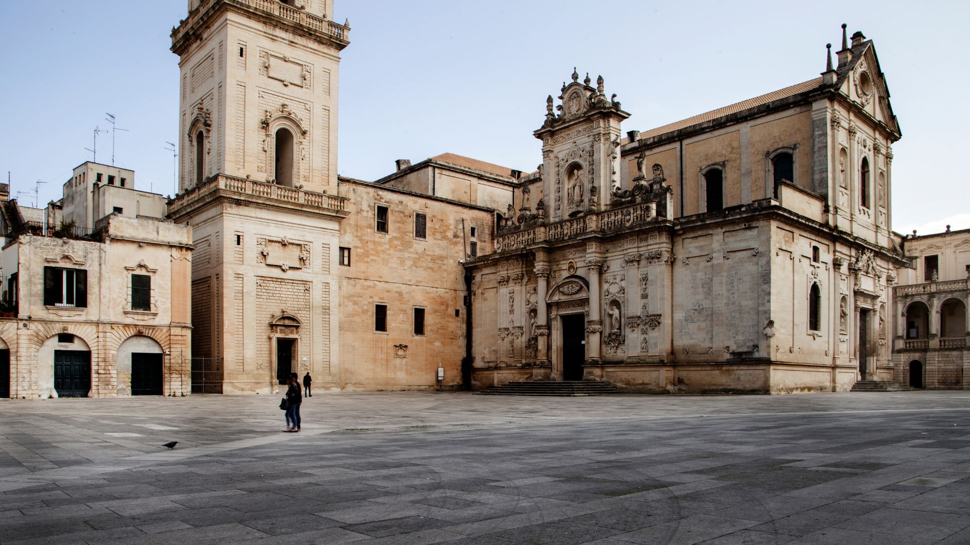 The Lecce Cathedral and its bell tower stand in a spacious, paved piazza under a clear sky.