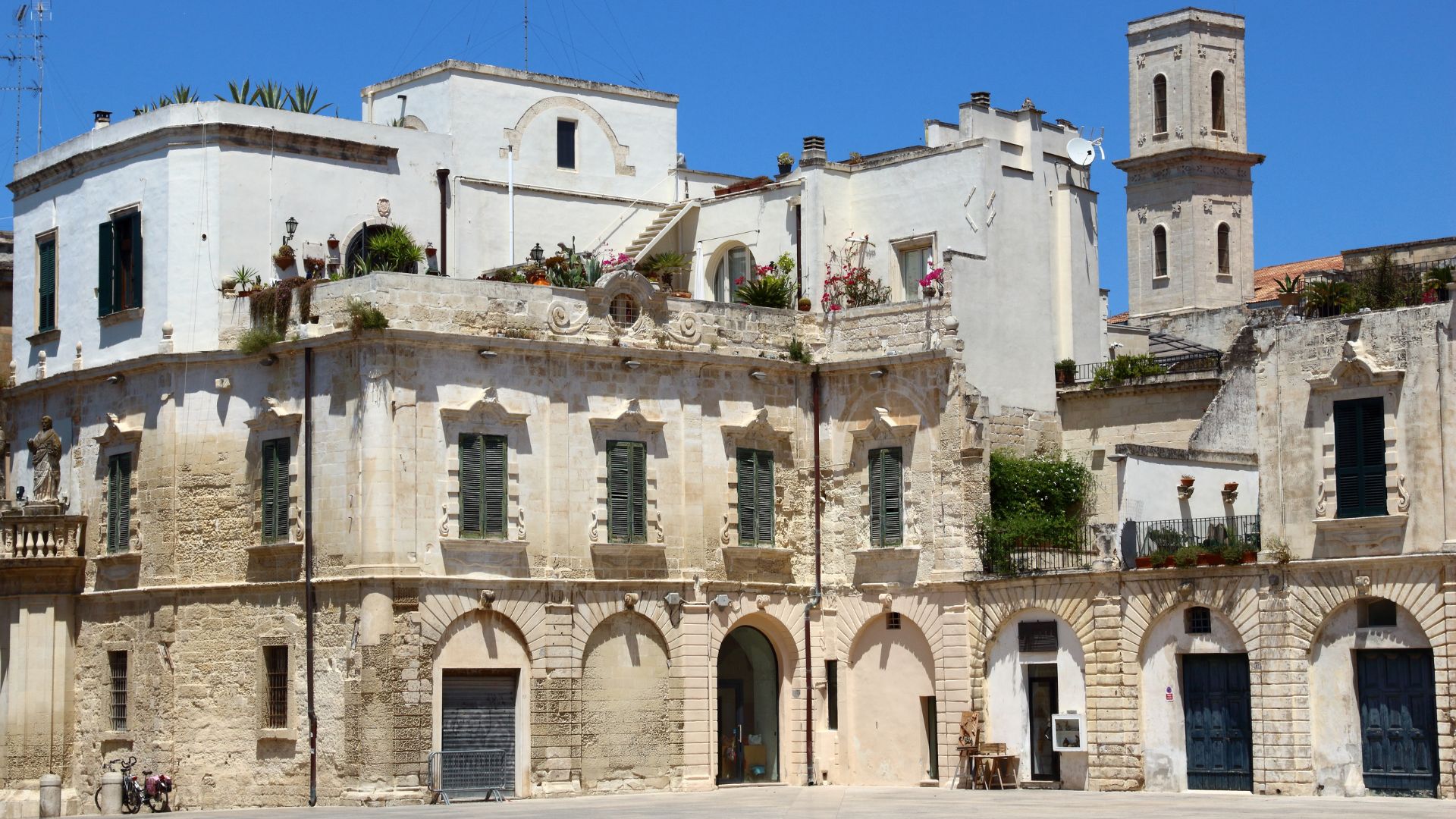 A multi-level building with white and beige stone in Piazza Mercantile, Lecce, Italy, featuring arched entryways and shuttered windows."