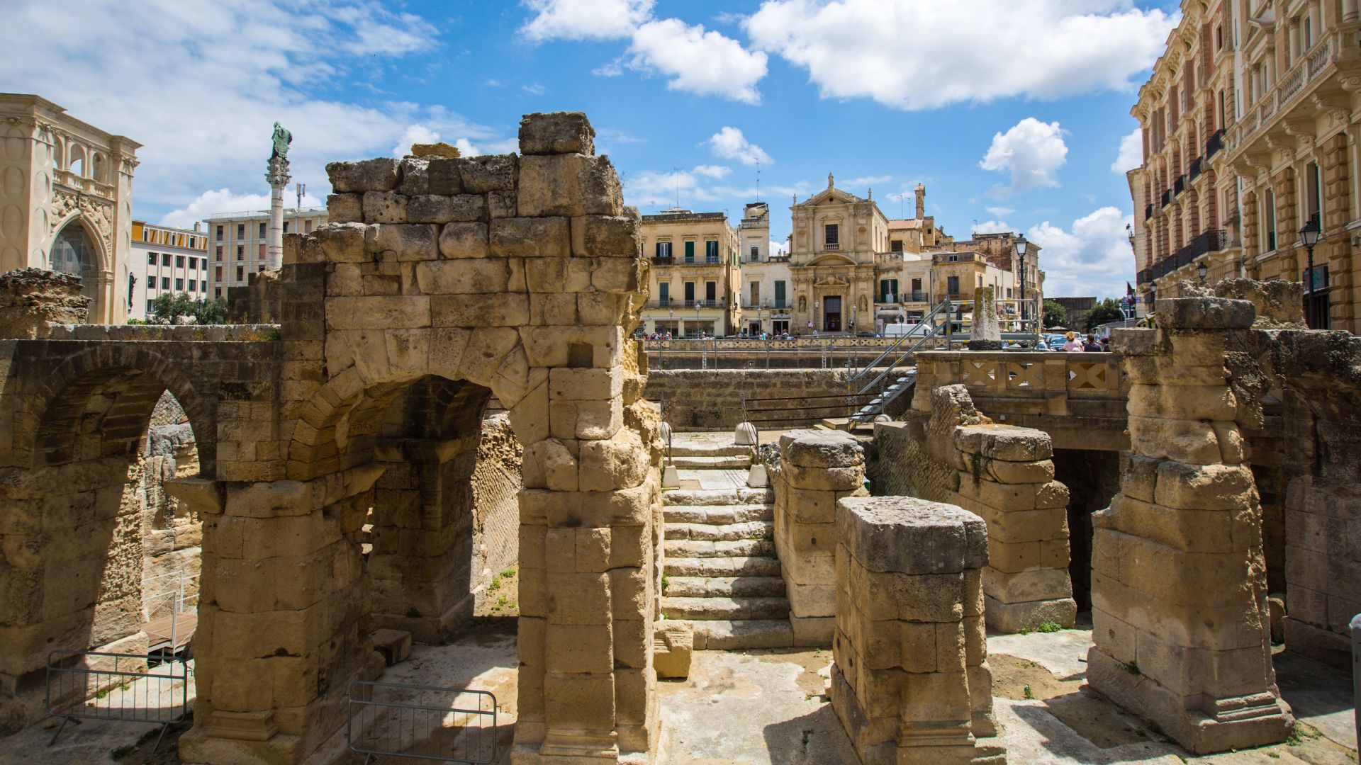The ruins of the ancient Roman Amphitheatre of Lecce, constructed from warm-toned stone blocks, dominate the foreground, with arched entryways and crumbling walls showcasing the structure's age. In the background, the cityscape of Lecce is visible, featuring buildings with similar stone coloring and a church with a dome, under a partly cloudy sky. 