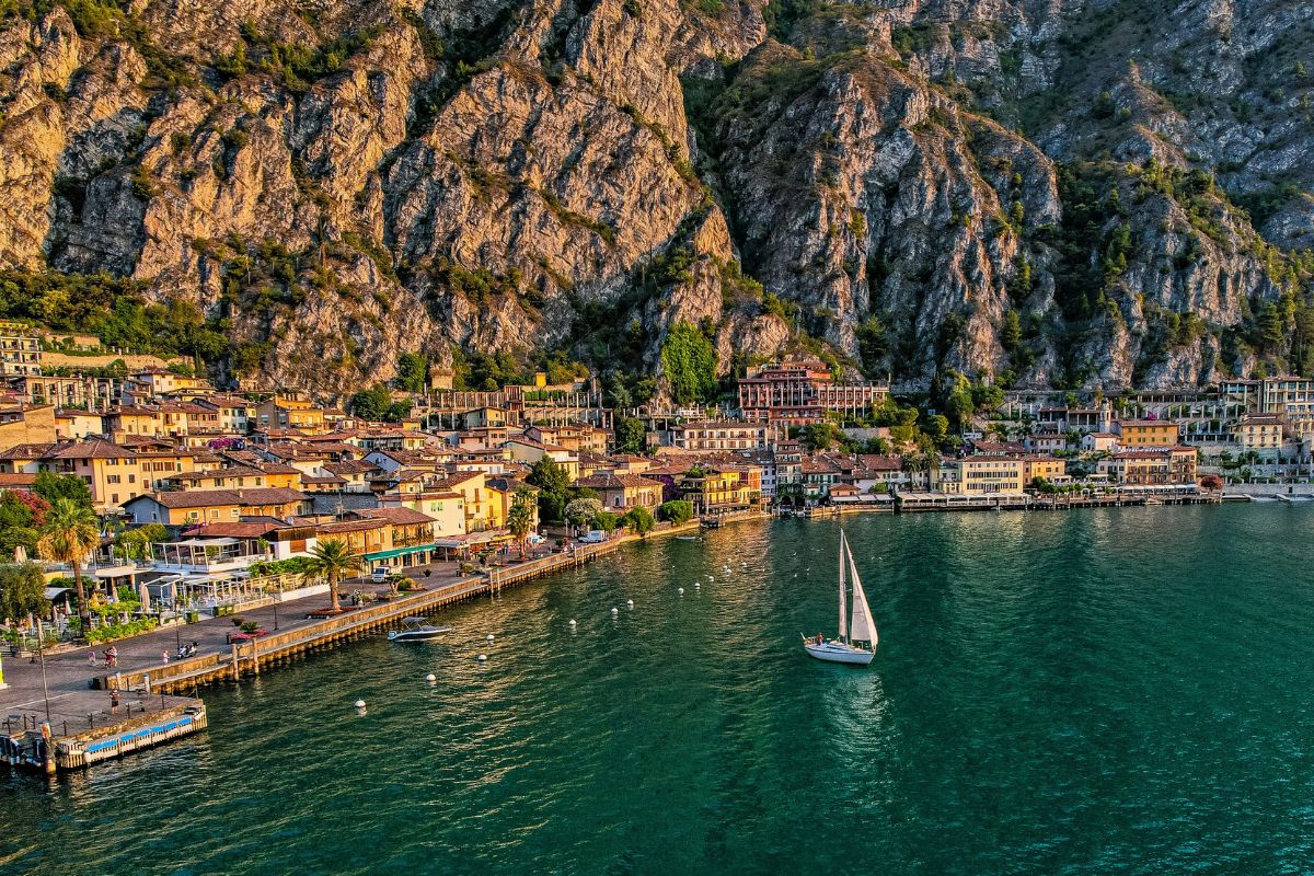 A landscape view of Lake Garda, featuring the town of Limone sul Garda nestled against rocky cliffs, with a sailboat on the lake's green waters. The town's buildings have a mix of orange and brown roofs, and the cliffs are rugged and textured. 