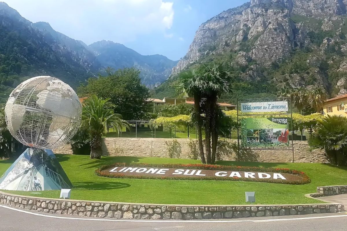 The image shows a scenic view of Limone Sul Garda, featuring a decorative globe sculpture on a roundabout, lush greenery, and mountains in the background. A sign reads "Welcome to Limone" in Italian. The town is nestled on the shores of Lake Garda in Italy, known for its lemon groves and picturesque landscapes. 