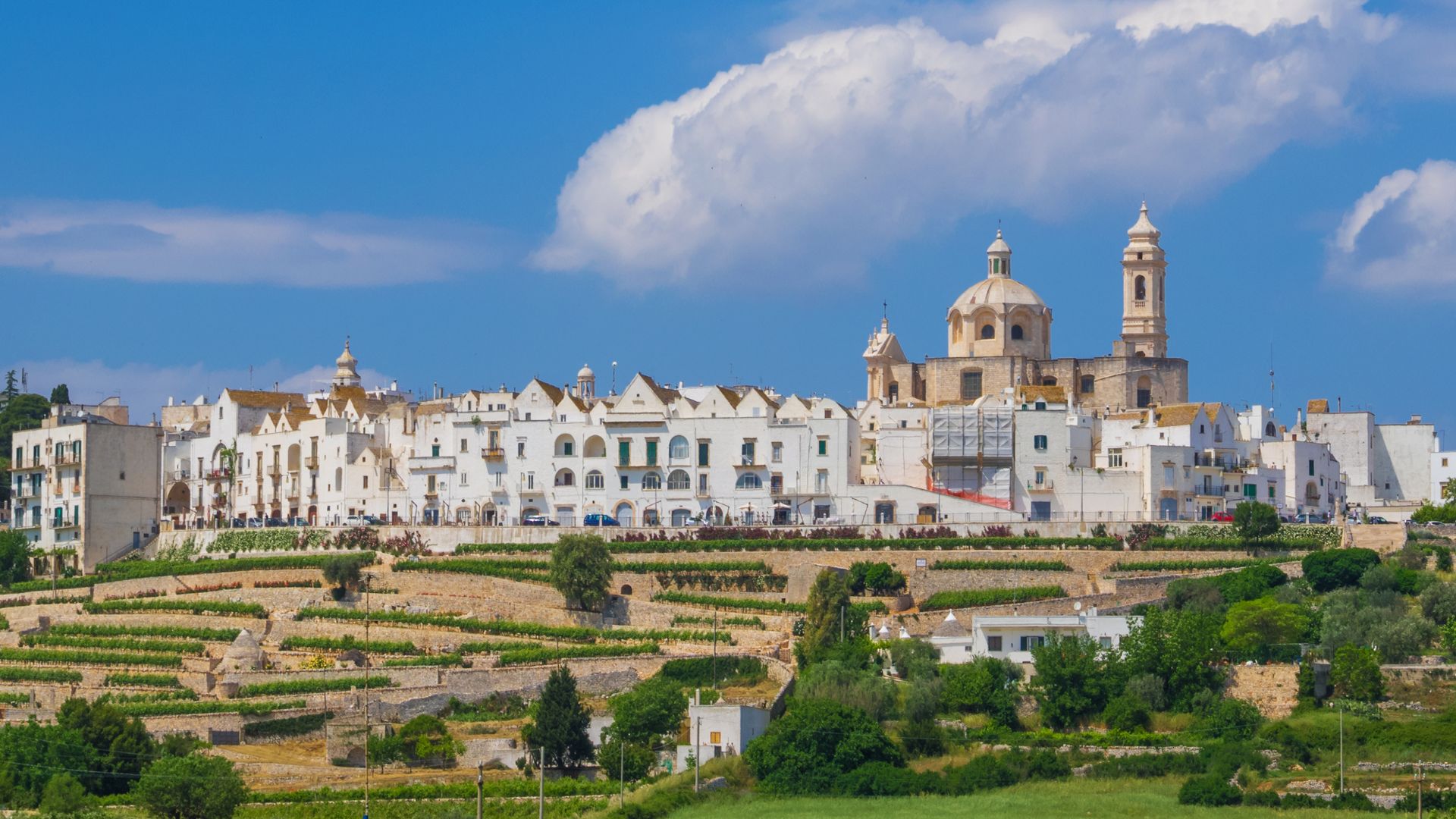 Locorotondo, a hilltop town in Italy's Itria Valley, is depicted with its whitewashed buildings and terraced landscape under a blue sky with clouds. The town's distinctive circular layout and the church's dome are visible, set against the backdrop of the valley's agricultural terraces.