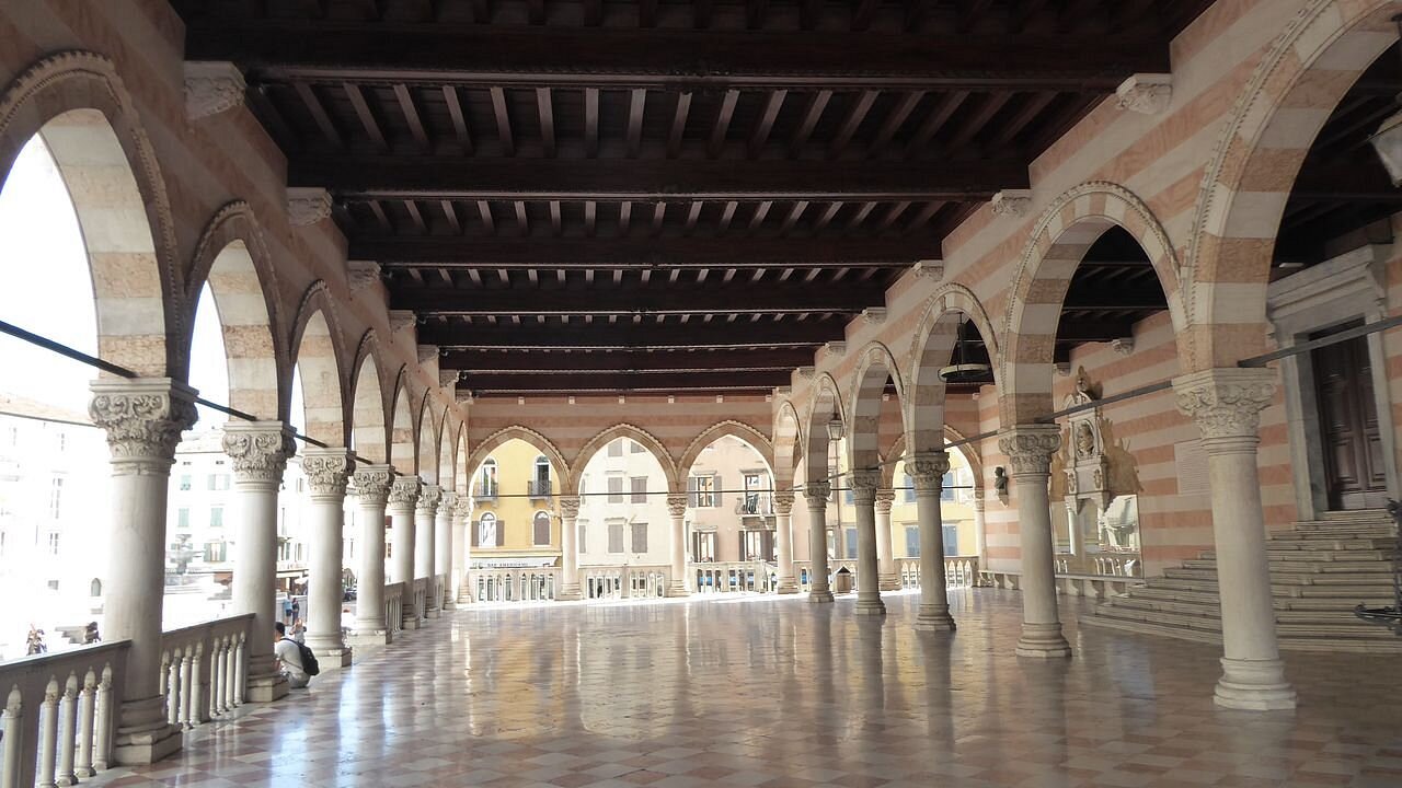 Interior view of the Loggia del Lionello in Udine, Italy, featuring elegant arches, striped walls, marble columns, and a polished checkered floor with a view of the square outside.