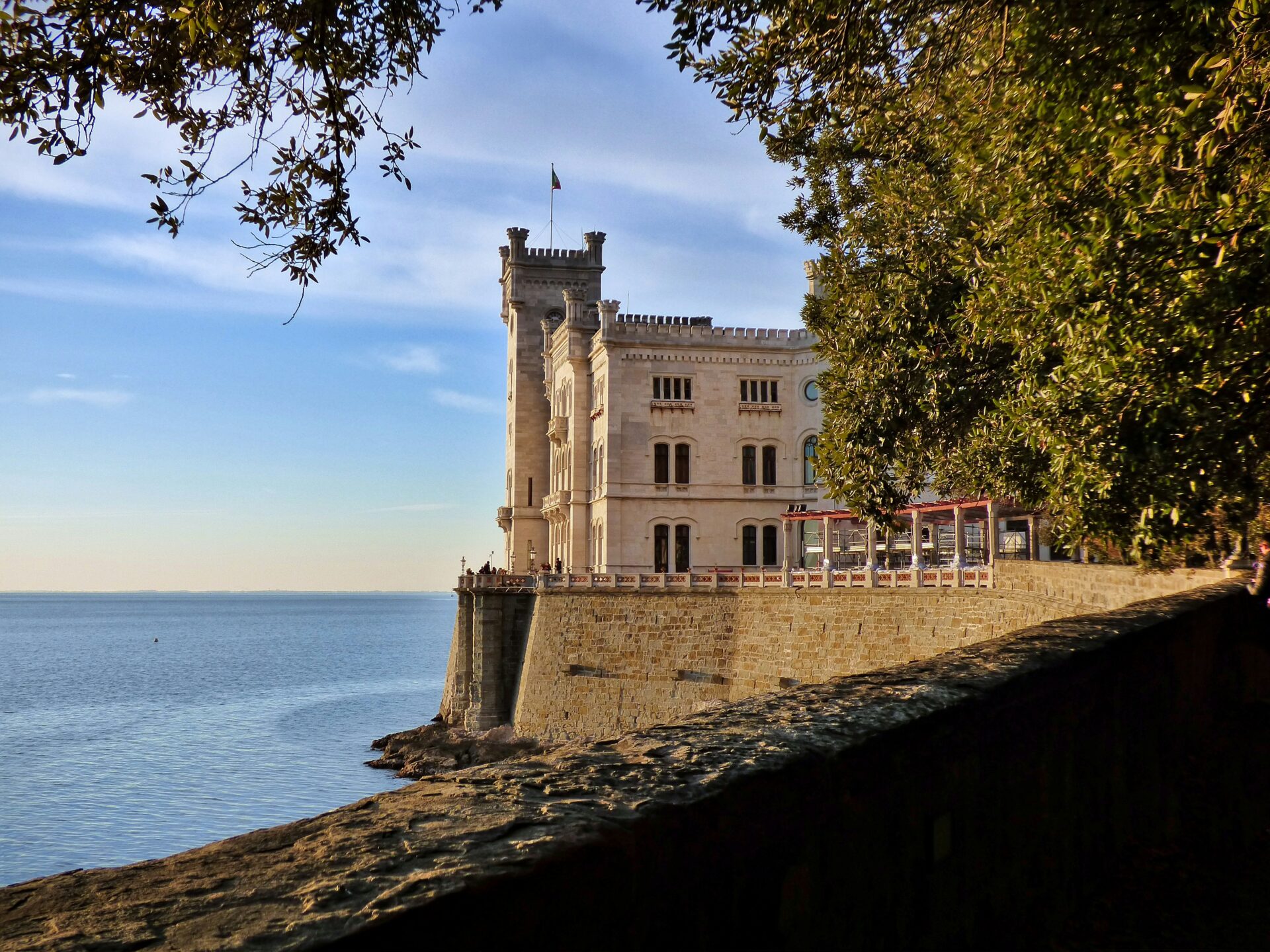 Miramare Castle in Trieste, Italy, perched on a rocky coastline overlooking the calm blue sea, framed by leafy trees in the foreground.