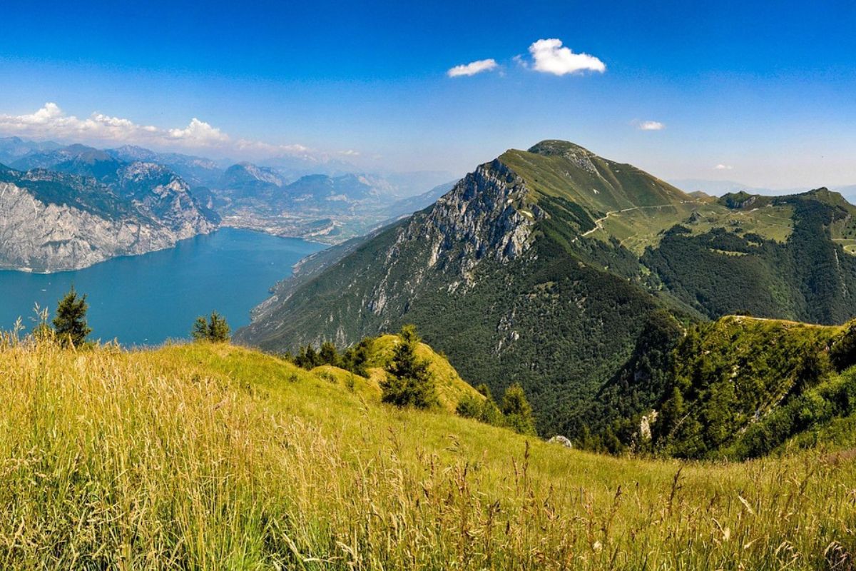 The image showcases the scenic landscape of Monte Baldo, likely situated near Lake Garda in Italy, with a clear blue sky dotted with a few clouds overhead. The foreground features grassy terrain, while the midground is dominated by the slopes of Monte Baldo, partially covered in lush greenery and rocky outcrops. In the distance, a large body of water, possibly Lake Garda, is visible, bordered by more mountains.