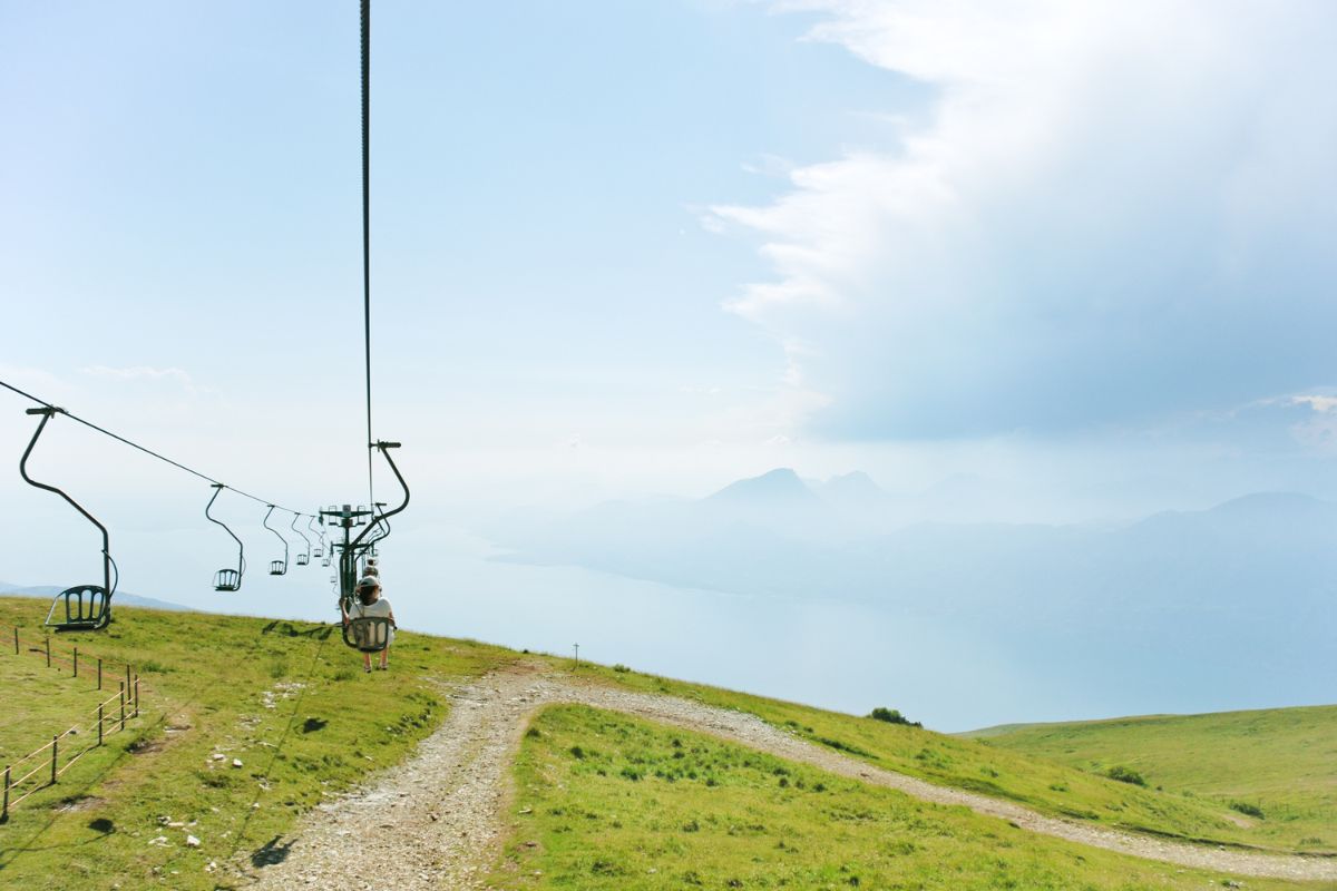 A chairlift ascends Monte Baldo, with a view of Lake Garda and the surrounding landscape in the background. The slopes of Monte Baldo are green, and the sky is partly cloudy.