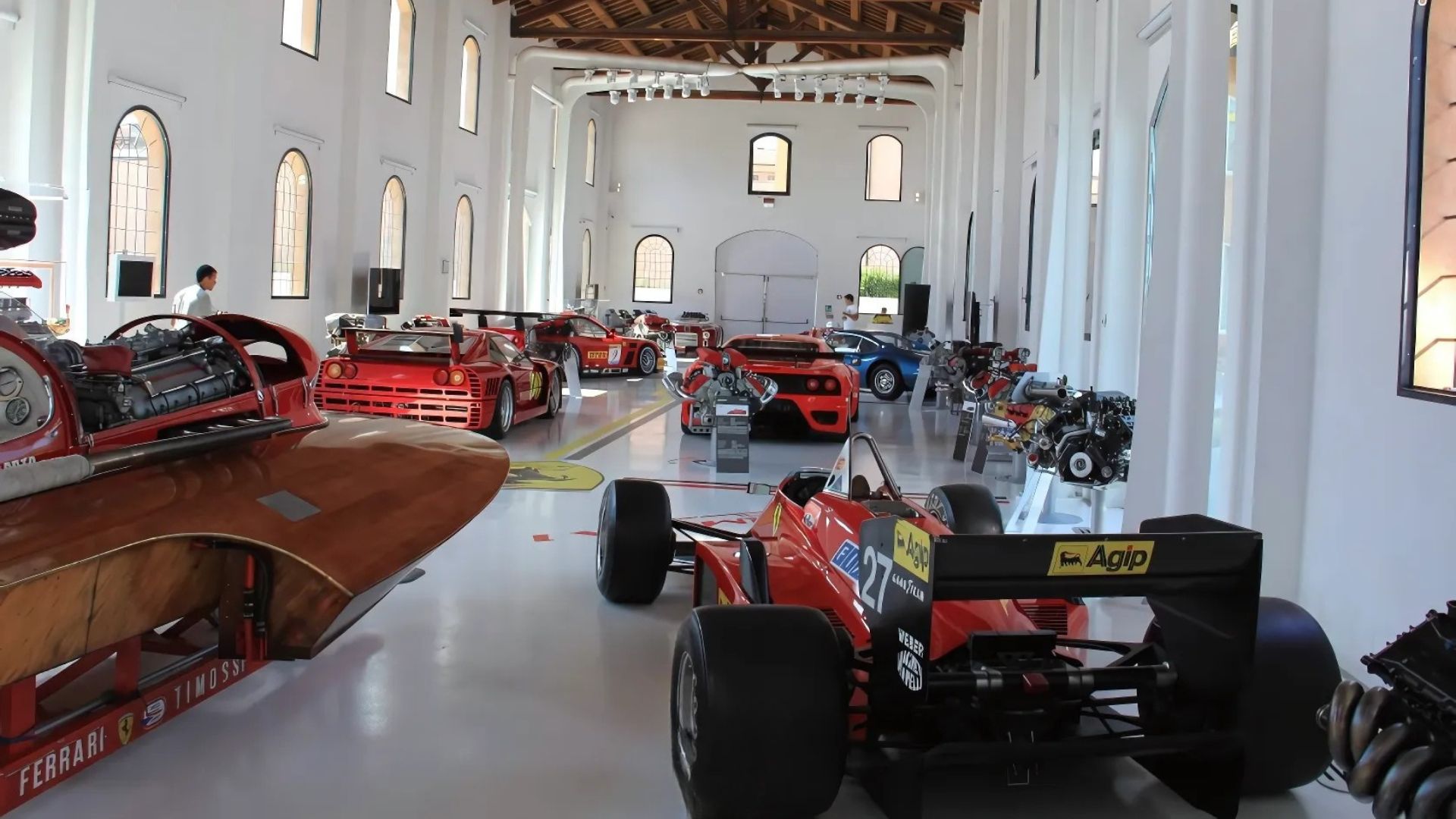 The scene is set in the Museo Enzo Ferrari in Modena, Italy, showcasing collection of classic Ferrari race cars and engines.