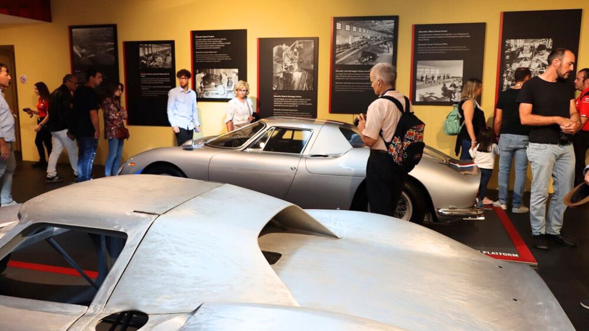 A silver Ferrari 250 LM is on display at the Museo Ferrari, surrounded by visitors and informational panels. 