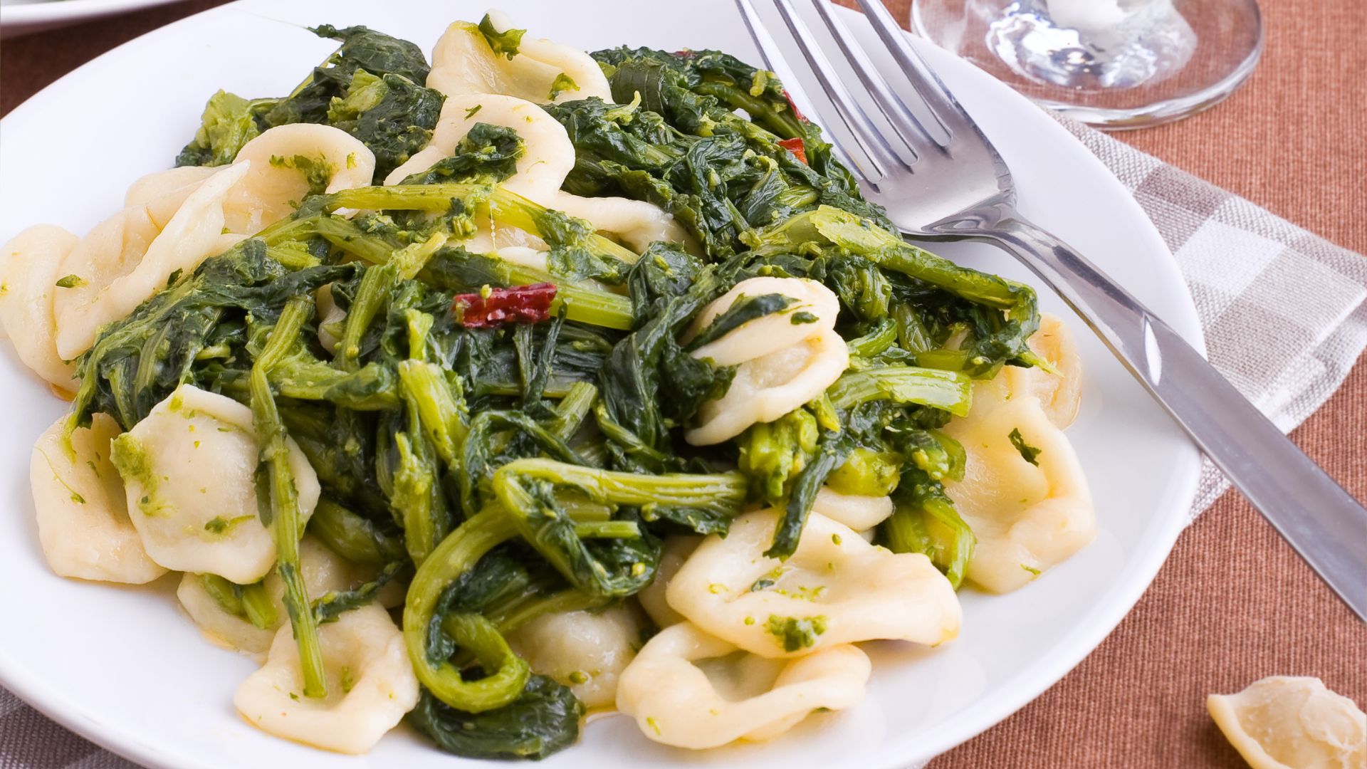 Orecchiette pasta with turnip tops is shown on a white plate, garnished with red pepper flakes and served with a fork.