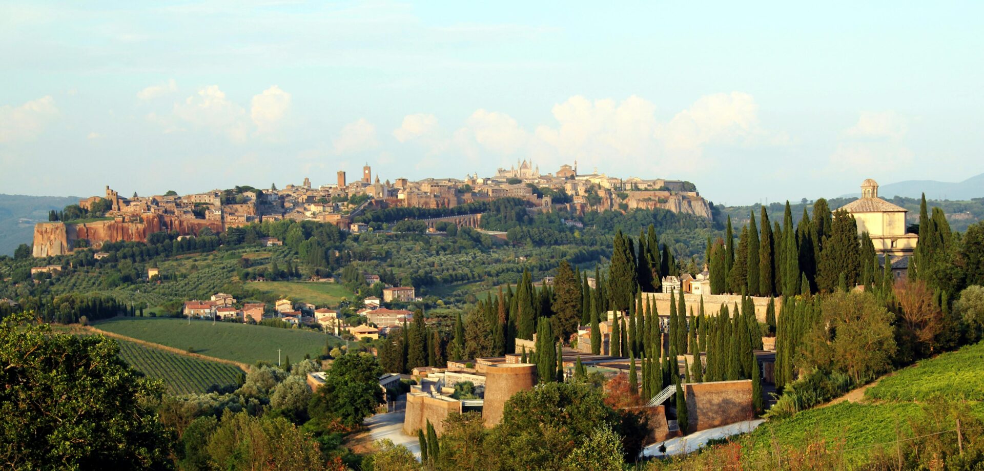 Panoramic landscape of Orvieto, Italy, featuring its cliffside medieval architecture, rolling green hills, and a backdrop of open countryside under a partly cloudy sky.