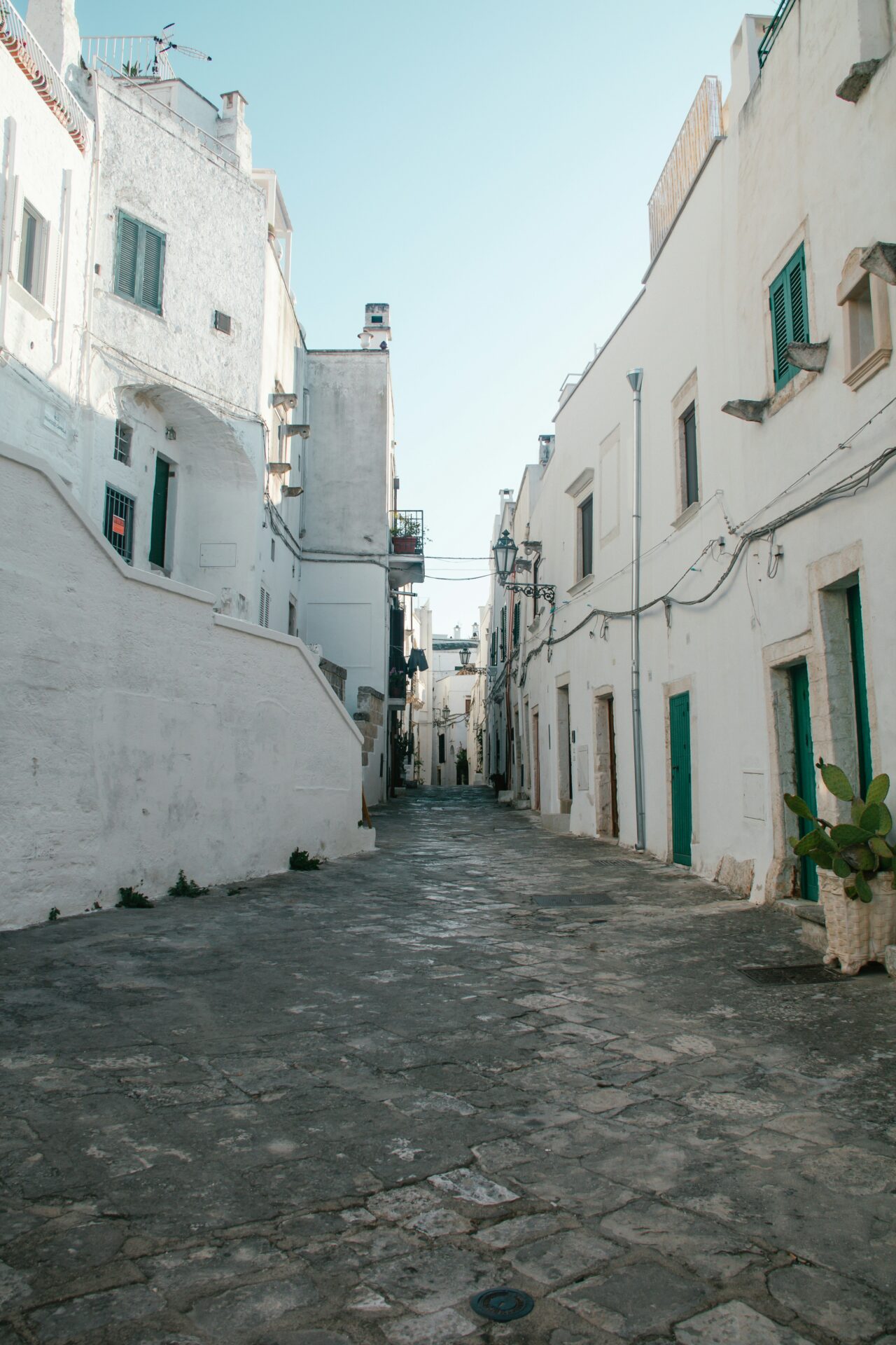 Narrow alleyway in Ostuni, Italy, lined with whitewashed walls, arched doorways, potted plants, and stone steps leading through the charming historic town.