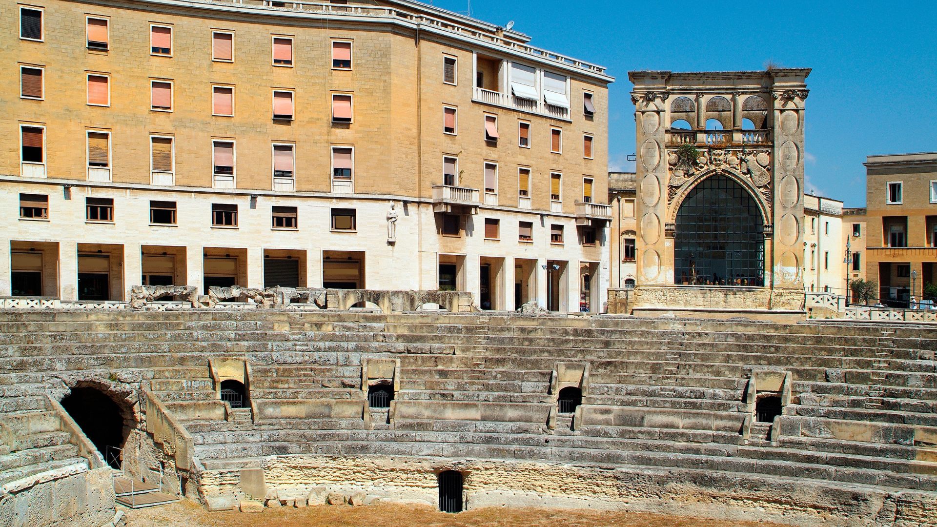 The image shows the Roman Amphitheatre of Lecce, situated in Lecce, Italy. The amphitheater is partially excavated and surrounded by modern buildings, including the Palazzo dell'Istituto Nazionale delle Assicurazioni and Palazzo del Seggio