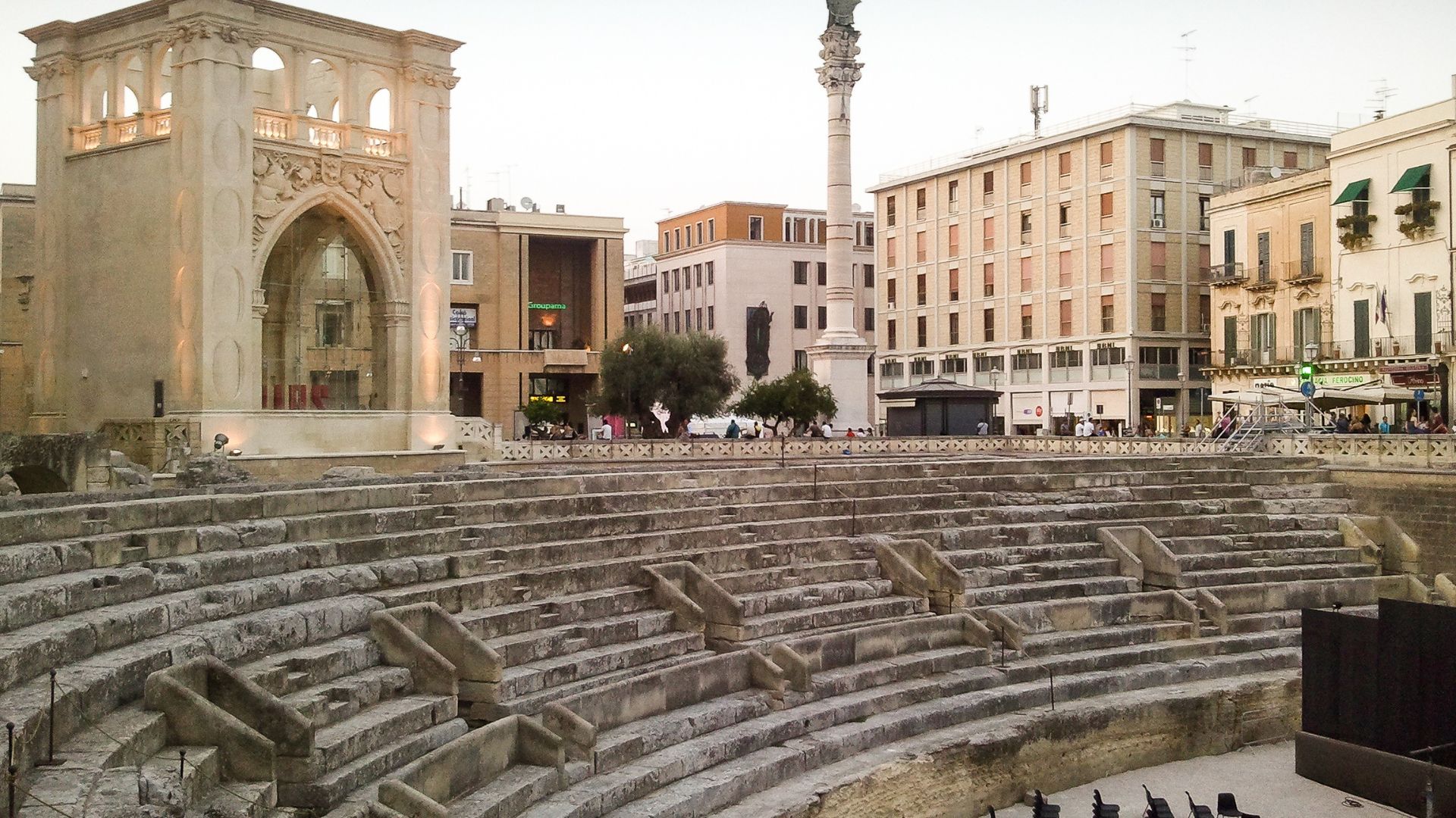 The image shows the Roman Amphitheatre of Lecce, situated in Lecce, Italy. The amphitheater is partially excavated and surrounded by modern buildings, including the Palazzo dell'Istituto Nazionale delle Assicurazioni and Palazzo del Seggio