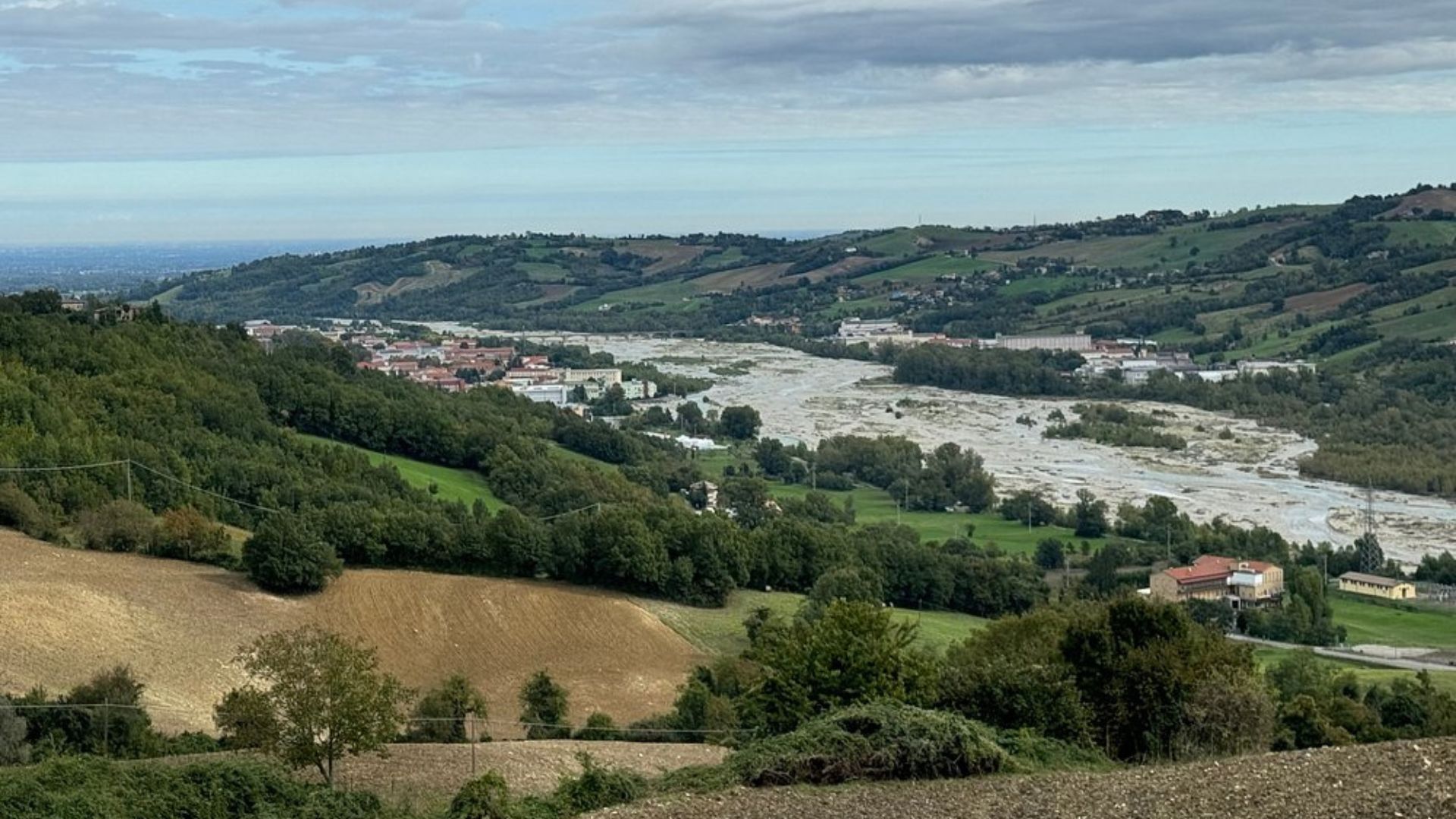 The image shows a landscape with a river or wide stream cutting through a valley, surrounded by hills and some buildings in Parma, Italy.
