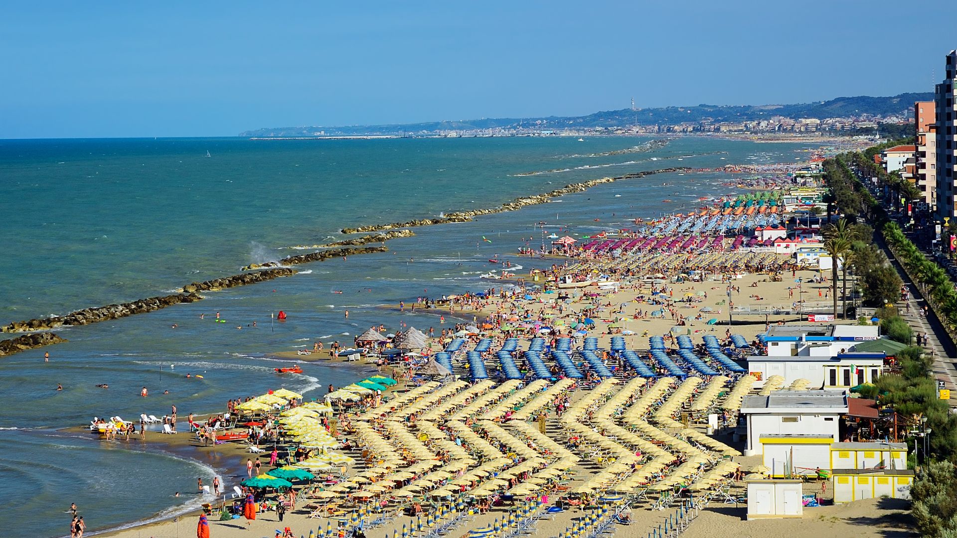 Image shows a colorful umbrellas, chairs, tables, and beach going on a beach in Trabocchi in Pescara, Italy.