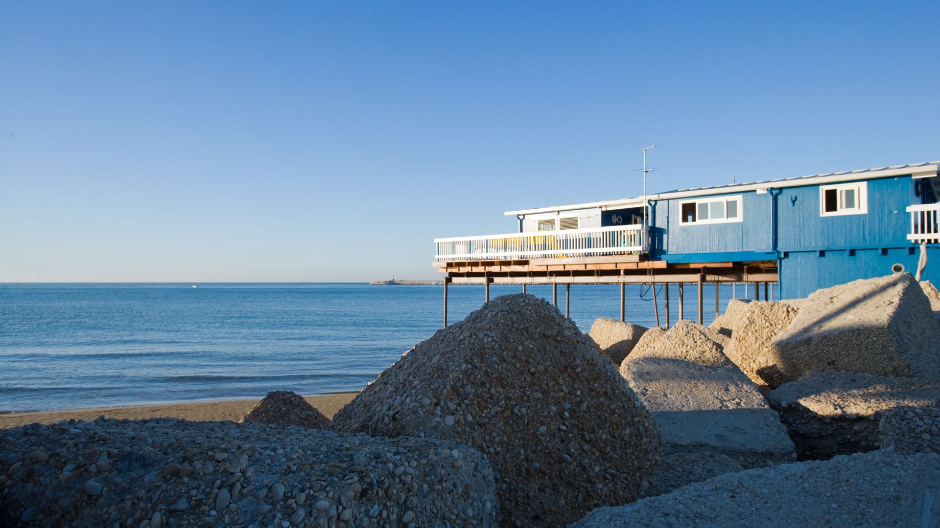 The image shows a building on stilts by the sea, with breakwater rocks in the foreground, which is consistent with the coastal setting of Pescara.