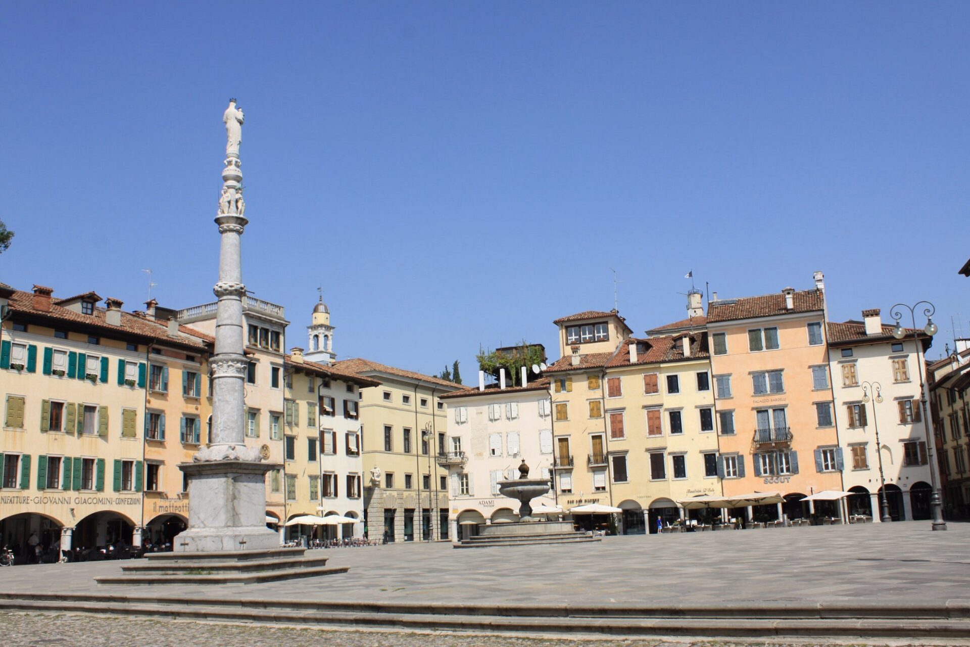 Bright and open view of Piazza San Giacomo in Udine, Italy, featuring a tall marble column, central fountain, and colorful historic buildings with shuttered windows under a clear blue sky.
