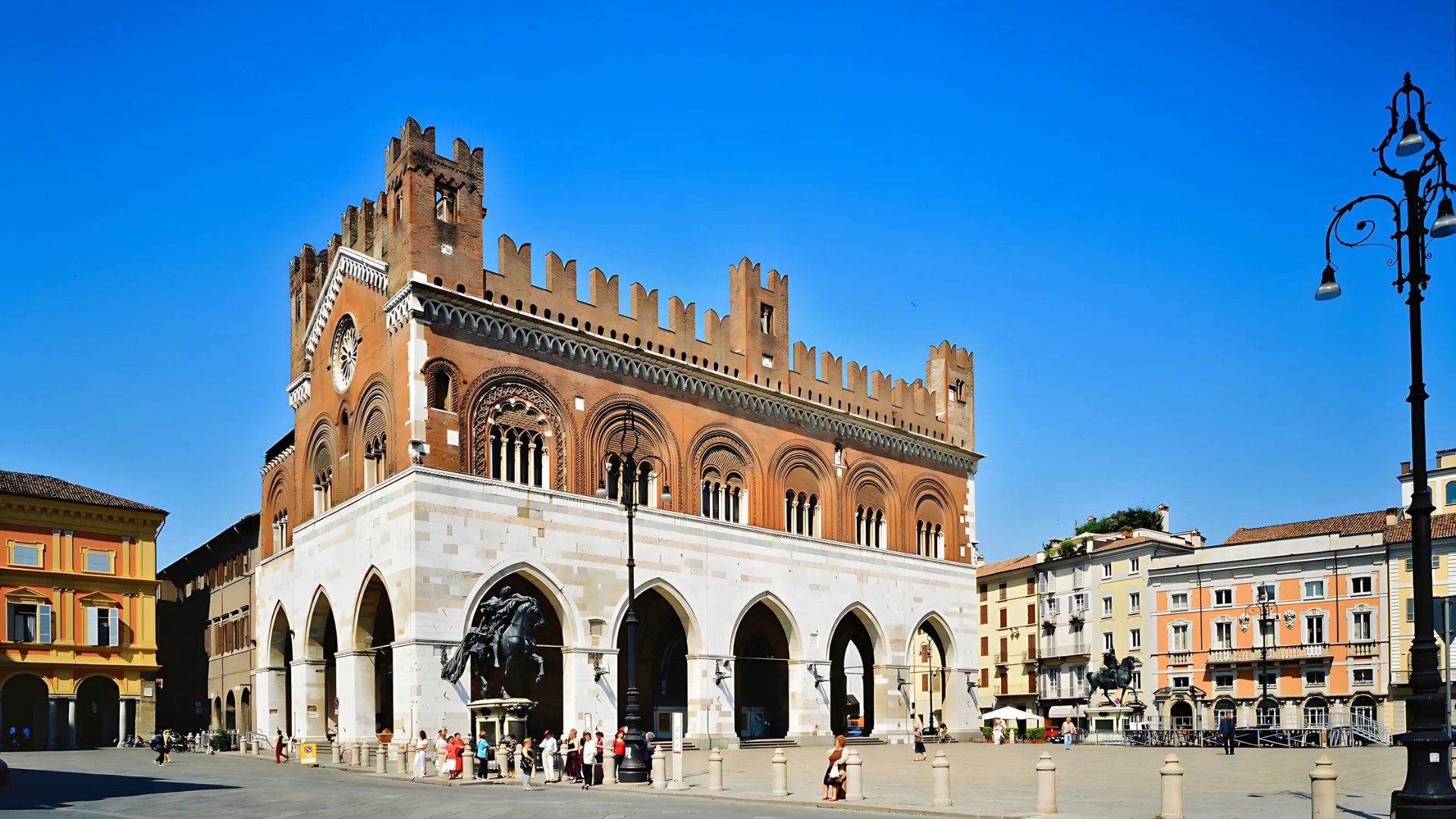 Piazza dei Cavalli in Piacenza, Italy, showcases the Palazzo Gotico with its distinctive Gothic architecture, including arched openings and crenellations, and equestrian statues in the foreground, all set against a clear blue sky. The surrounding buildings and people in the piazza suggest a lively urban environment.