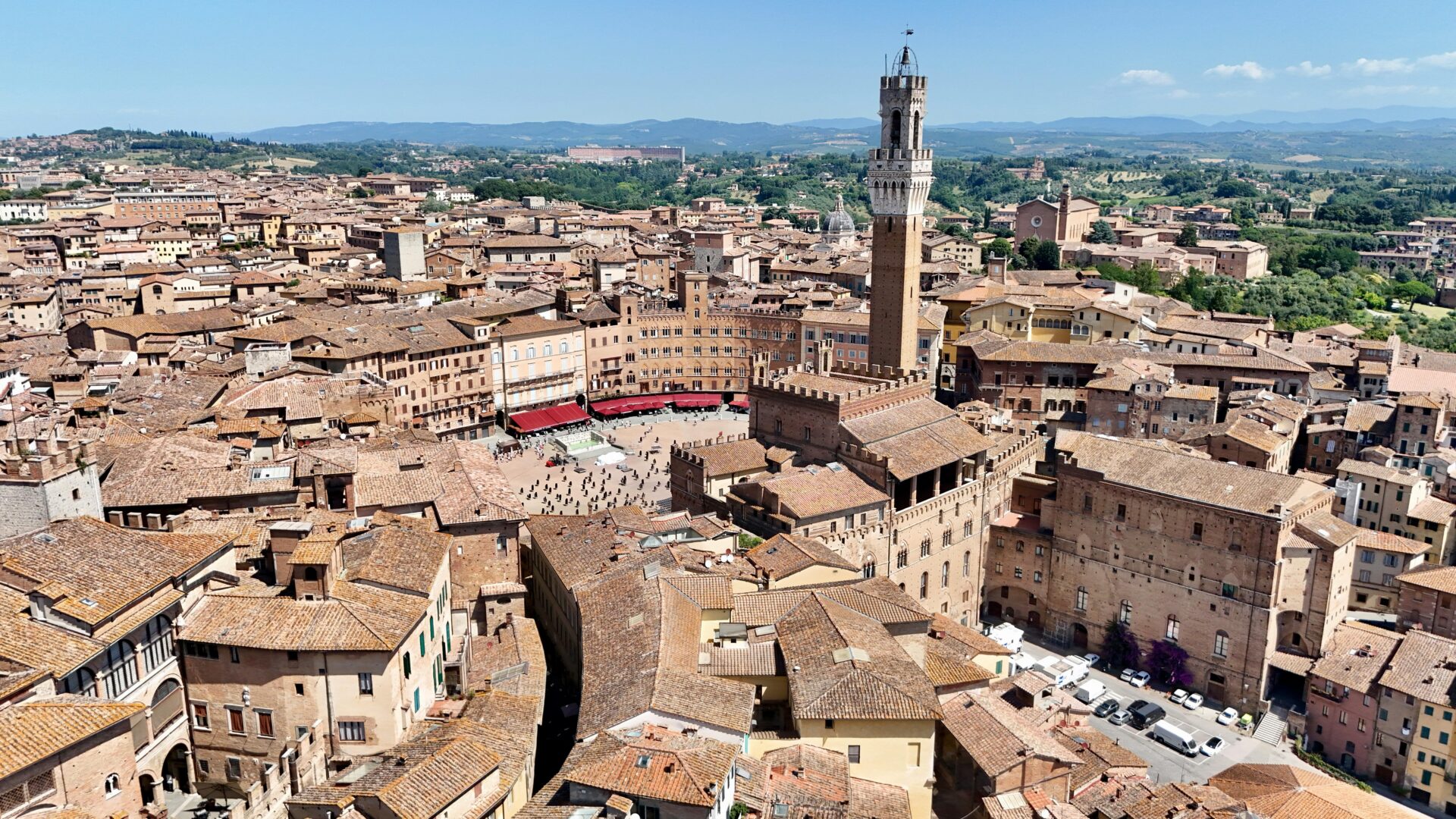 Piazza del Campo in Siena, Italy, a spacious fan-shaped square lined with historic buildings and cafes, featuring the Palazzo Pubblico and Torre del Mangia under a sunny sky
