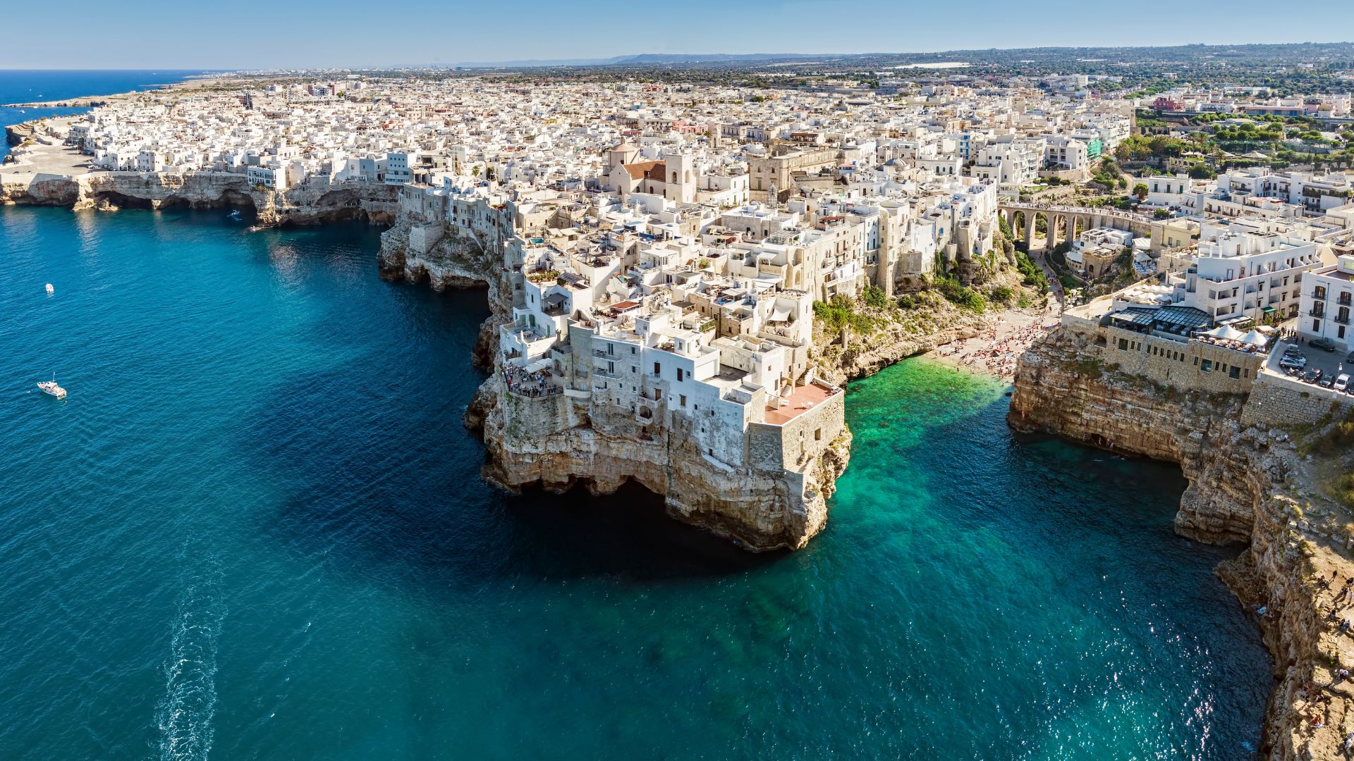 Image shows the clear turquoise water, cliff and the shore with some beach goers in Polignano a Mare.