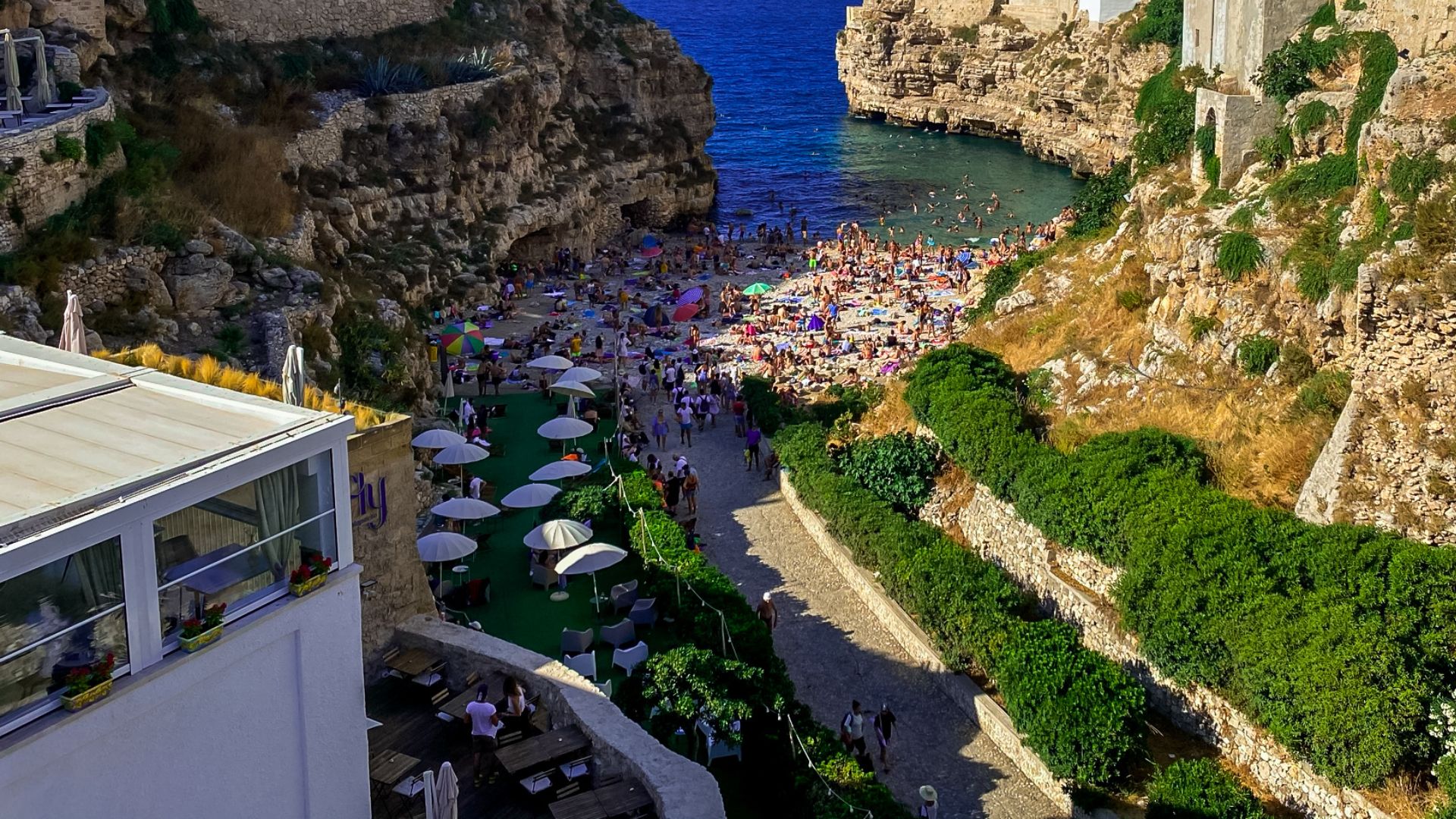 Image shows an aerial view of the road to the shore of the beach with the beach goers in Polignano a Mare. 