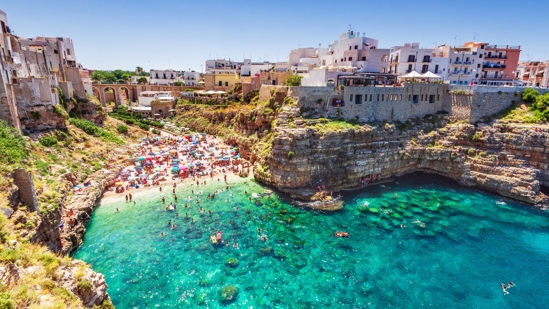 Image shows the clear turquoise water, cliff and the shore with some beach goers in Polignano a Mare, Italy. 