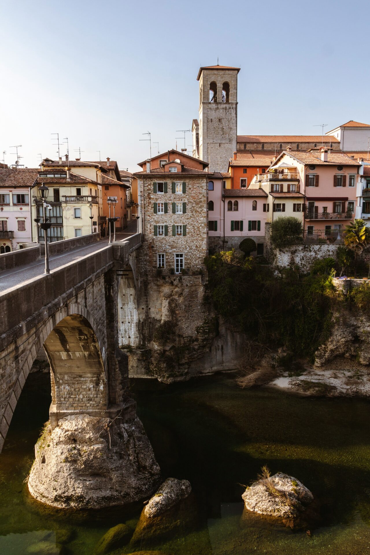 Ponte del Diavolo, a historic stone bridge in Cividale del Friuli, Italy, crossing over a clear river with colorful buildings and a bell tower in the background.