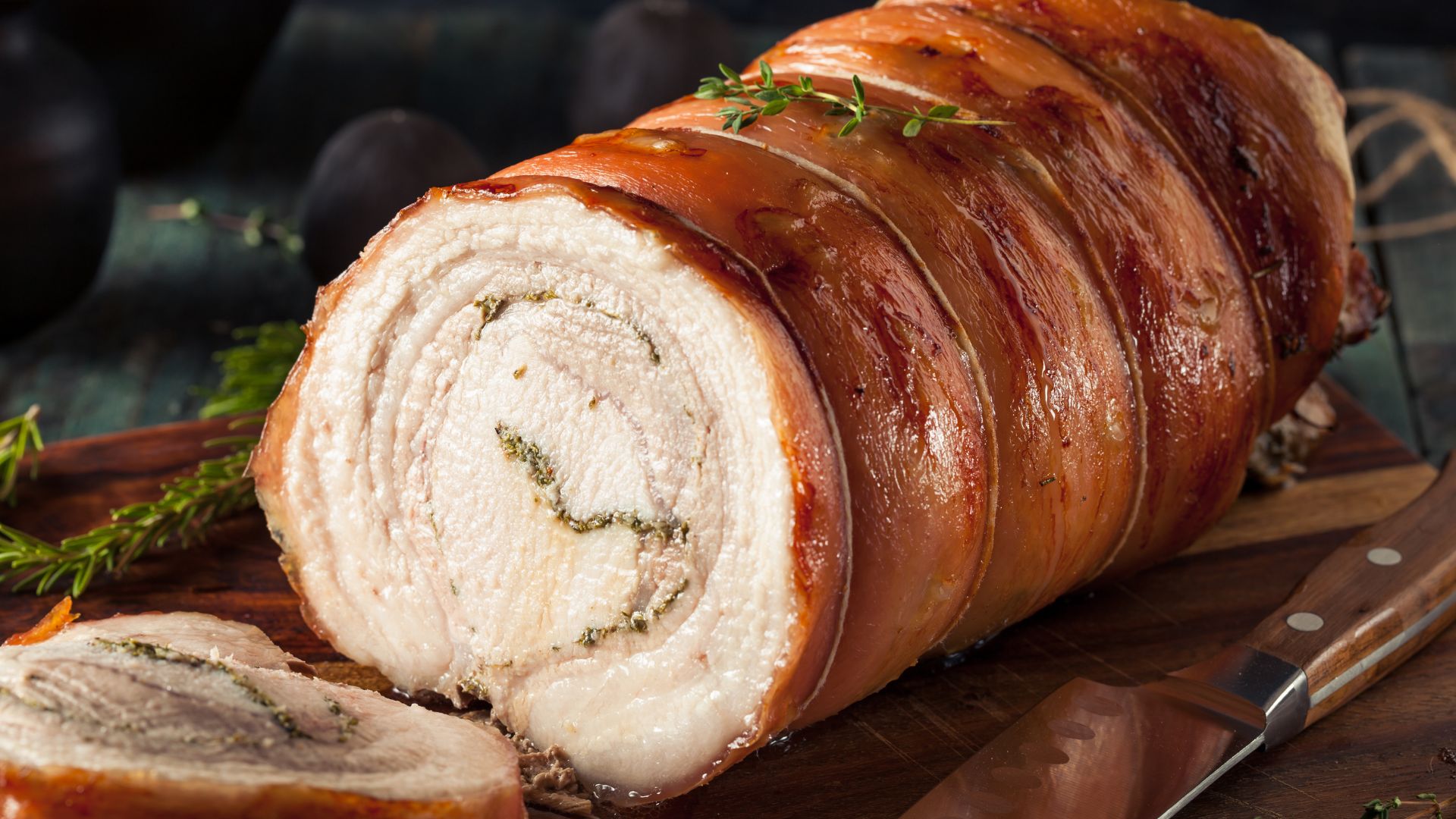 Image of Porchetta on a board with herbs and a knife.
