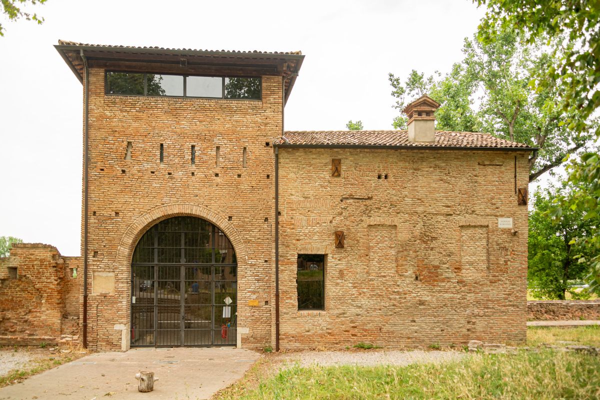 The image shows the Porta degli Angeli. It is a medieval gate constructed of brick, featuring a large arched entrance with a metal gate, smaller windows, and a tiled roof.